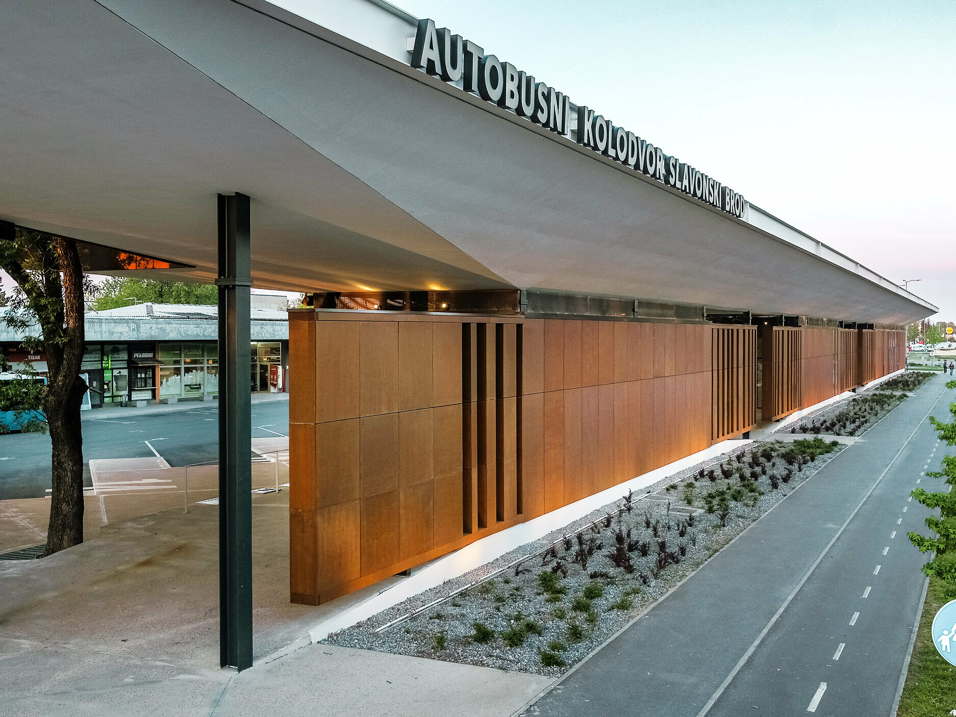 La photo montre le hall moderne de la gare routière « Autobusni Kolodvor Slavonski Brod » en Croatie. Le hall de gare présente une façade remarquable de panneaux bruns soutenus par des colonnes noires. Le toit Prefalz blanc de PREFA s'étire sur toute la structure, protégeant les espaces situés en dessous. Au premier plan, on peut voir une piste cyclable flanquée d'un espace vert bien entretenu et de jeunes arbres. À l'arrière-plan, on distingue un bus bleu clair, ainsi que divers magasins.