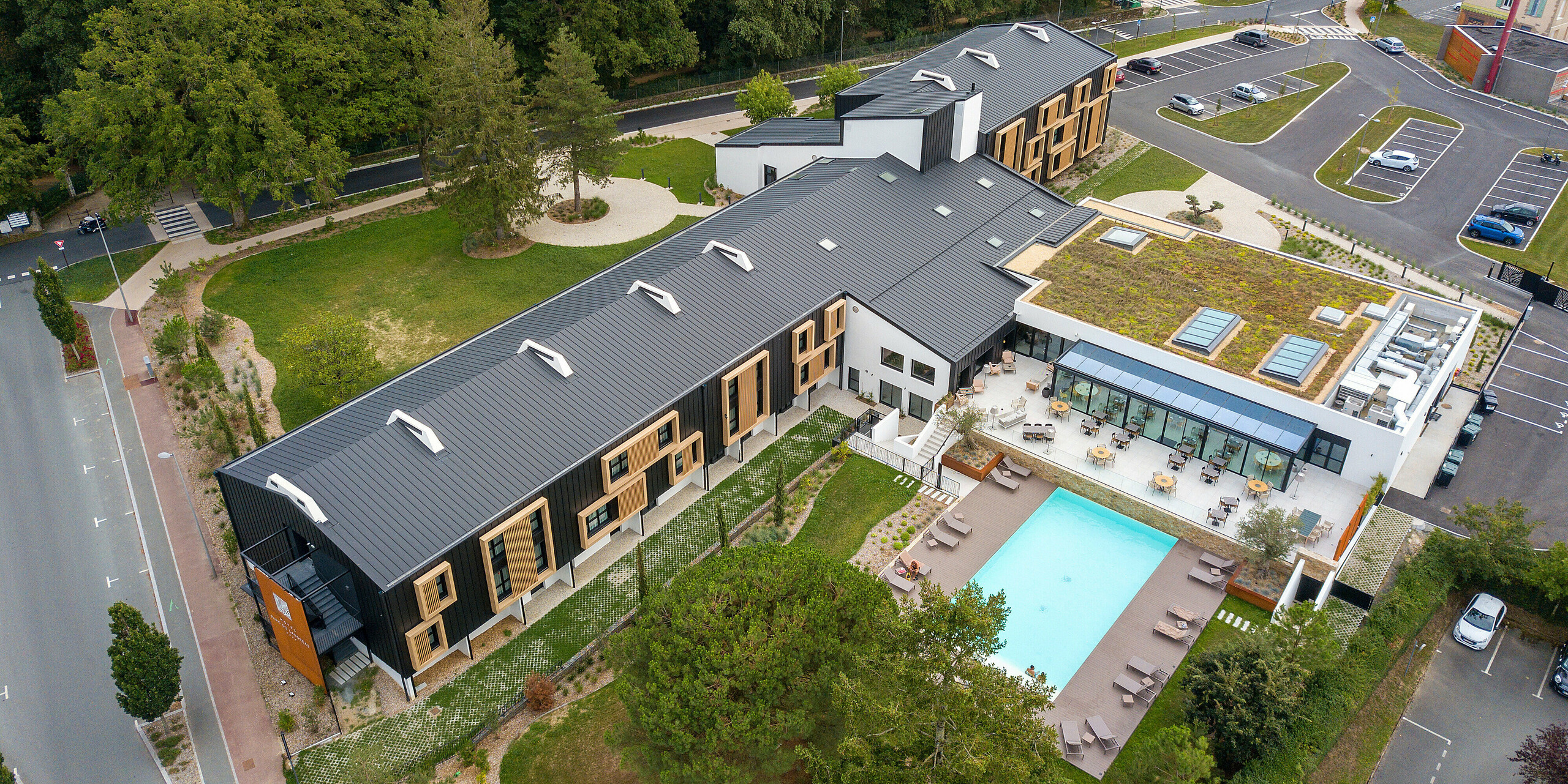 [Translate to Français:] Luftaufnahme der Rückseite des Hôtel Parc du Landreau in Les Herbiers, Frankreich, mit dem PREFALZ Dach- und Fassadensystem in Schwarzgrau. Naturbelassene Holzelemente um die Fenster unterstreichen das moderne Gestaltungskonzept. Ein Gebäudeteil wurde mit einem begrünten Dach ausgestattet, das die langlebige Aluminium-Stehfalzdeckung optisch ergänzt. Der großzügige Außenbereich mit Pool und Terrasse bietet den Gästen eine einladende Atmosphäre.