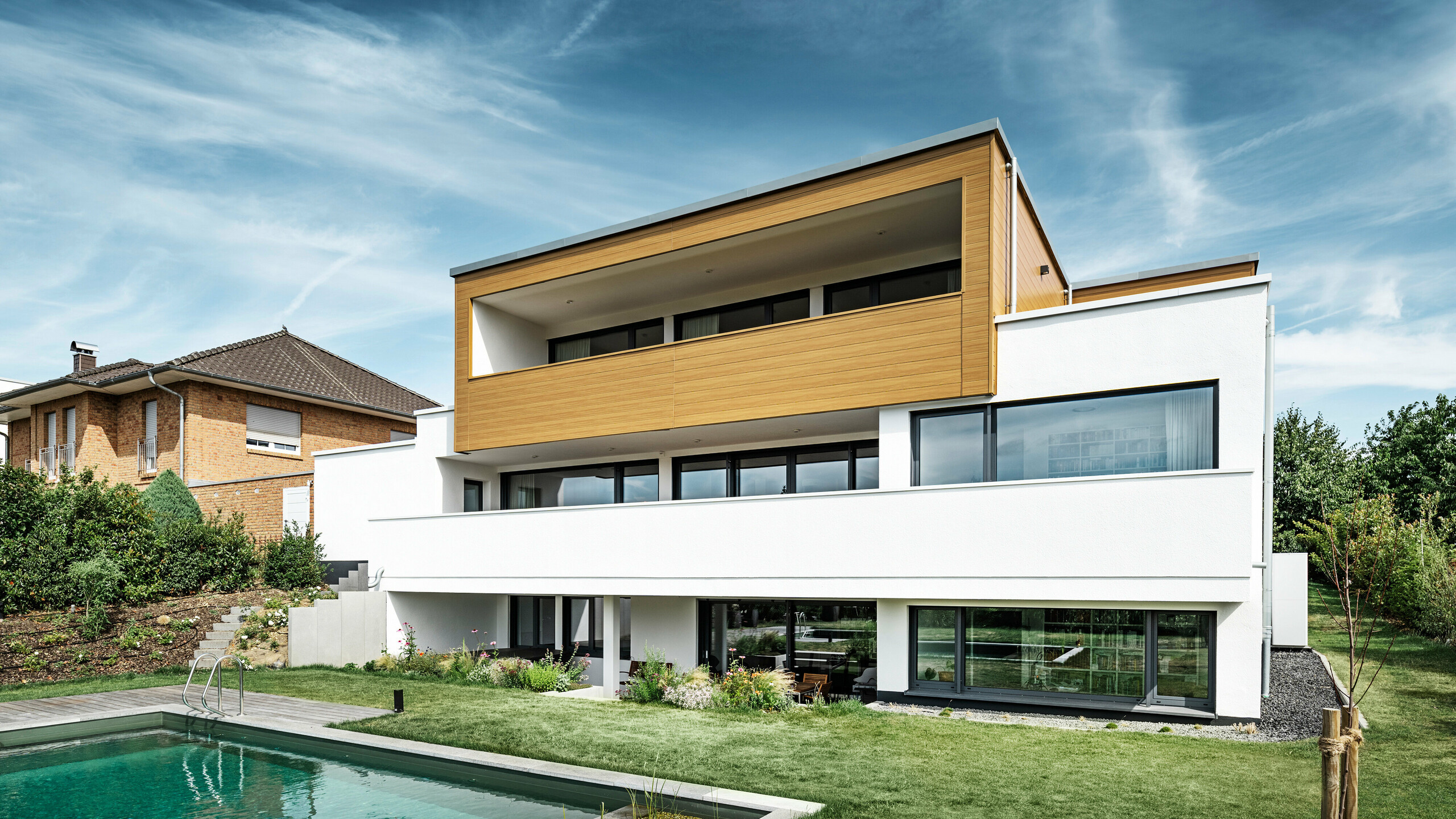 L'arrière d'une maison individuelle moderne à Usingen, Allemagne. En bas de l'image, on peut voir une pelouse verte, une piscine ainsi qu'un jeune arbre de jardin. Cette maison remarquable dispose d'un socle de bâtiment blanc composé d'une façade en crépi, d'une façade en aluminium à l'étage supérieur en PREFA Sidings en chêne naturel ainsi que de grandes surfaces de fenêtres avec un cadre de couleur anthracite. De loin, on distingue la maison voisine avec une façade en briques sur le bord gauche de l'image.