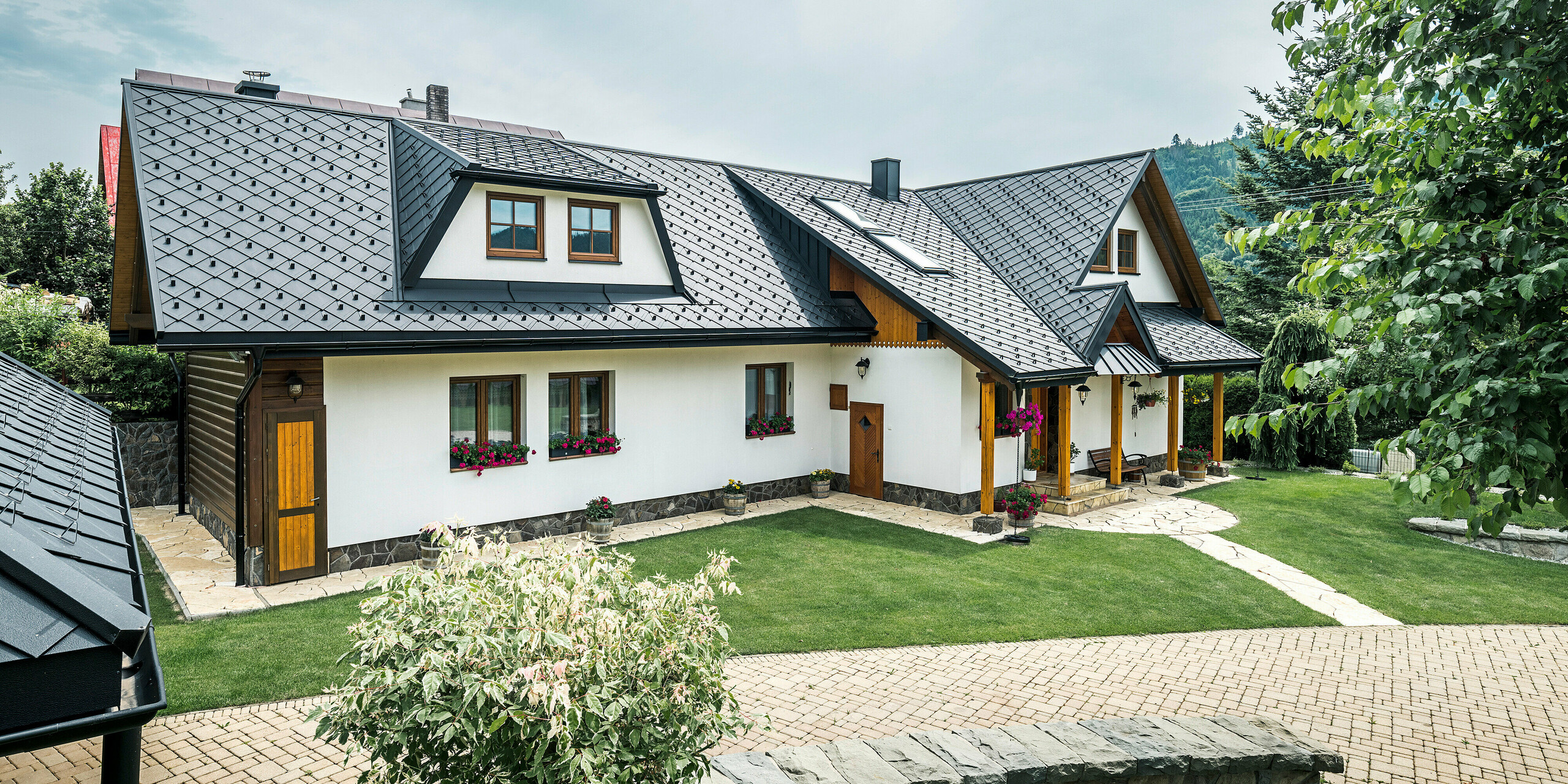 Vue d'une maison individuelle traditionnelle à Stara Bystrica, en Slovaquie, avec un toit en losanges PREFA 29 × 29 en P.10 noir. Les losanges en aluminium s'étendent également sur la lucarne trapézoïdale de la façade du bâtiment. Au premier plan de la photo, vous pouvez voir un jardin bien entretenu. La zone d'entrée est protégée par un auvent recouvert de PREFALZ. L'allée et les compositions florales soigneusement conçues soulignent l'image globale accueillante et élégante.