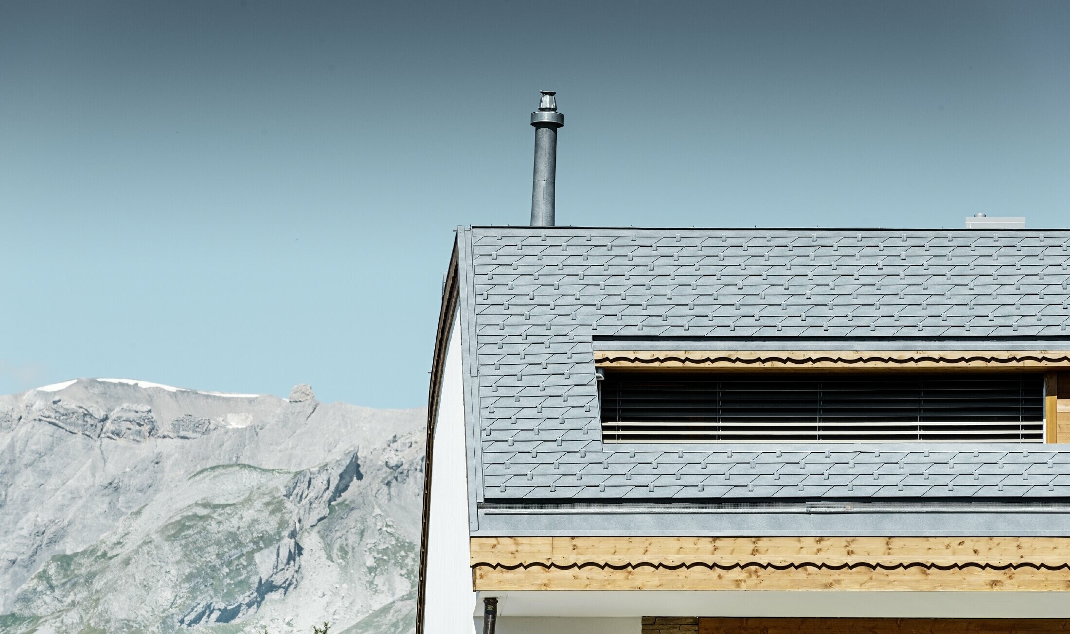 Immeuble d’habitation de Crans-Montana (Suisse) avec les Alpes en arrière-plan — Façade intégrant de nombreux éléments en bois et dont la toiture a été réalisée avec des bardeaux en aluminium PREFA de couleur gris pierre