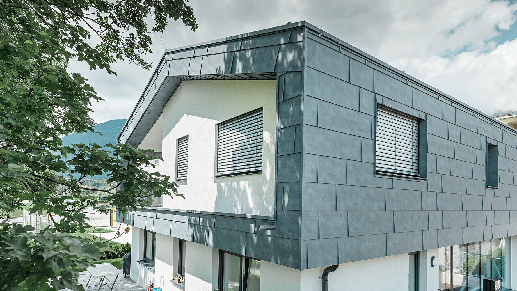 Maison individuelle moderne sans avancée de toit. L’étage supérieur a été recouvert de panneaux de façade FX.12 PREFA couleur gris pierre et le reste de la façade a été crépi en blanc.