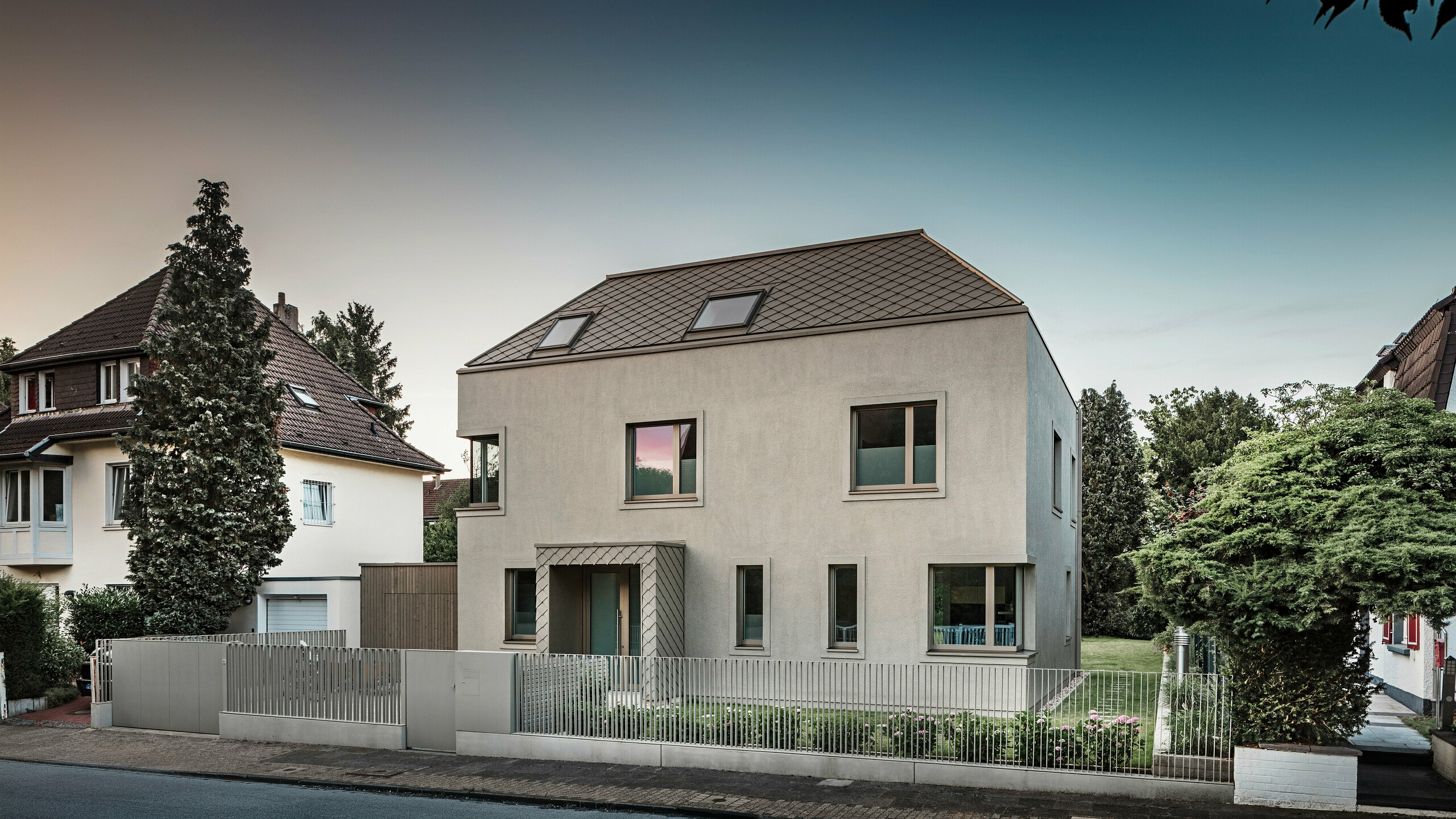 Une maison individuelle moderne avec un toit en croupe et un revêtement de toiture et de façade en aluminium couleur bronze de PREFA. La maison est située dans un quartier résidentiel calme, entourée de jardins bien entretenus et d'autres maisons individuelles. Au premier plan, vous pouvez voir une clôture basse qui sépare la propriété de la rue. L'architecture de la maison se caractérise par des lignes épurées et de grandes fenêtres qui laissent entrer beaucoup de lumière naturelle dans les intérieurs.