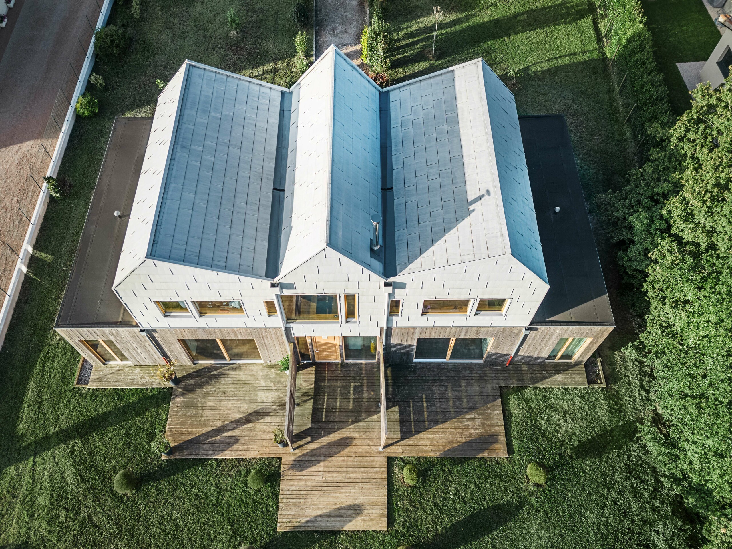 Vue plongeante d'une maison jumelée avec un toit à pignon en aluminium de PREFA. Le toit présente des lignes épurées et une finition moderne. La terrasse en bois complète le caractère moderne de la maison.