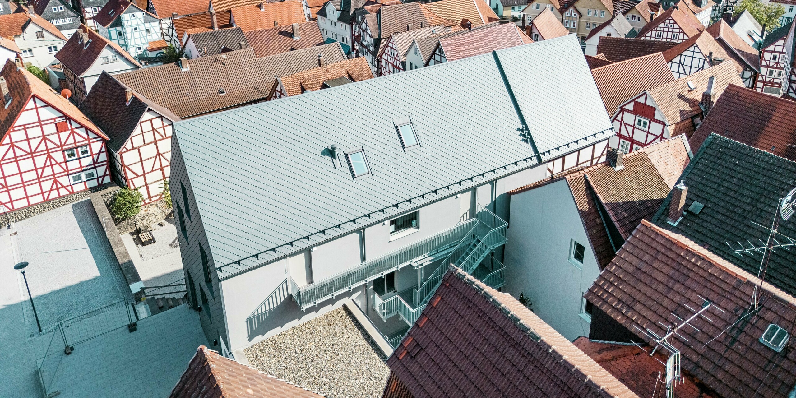 Vue grand angle par drone du Volkshaus à Homberg, DE, avec des losanges de toiture 29x29 PREFA en P.10 gris pierre. La conception moderne du toit en aluminium robuste s'intègre harmonieusement dans la vieille ville historique avec ses toits de tuiles rouges et offre durabilité, protection contre les intempéries et élégance intemporelle.