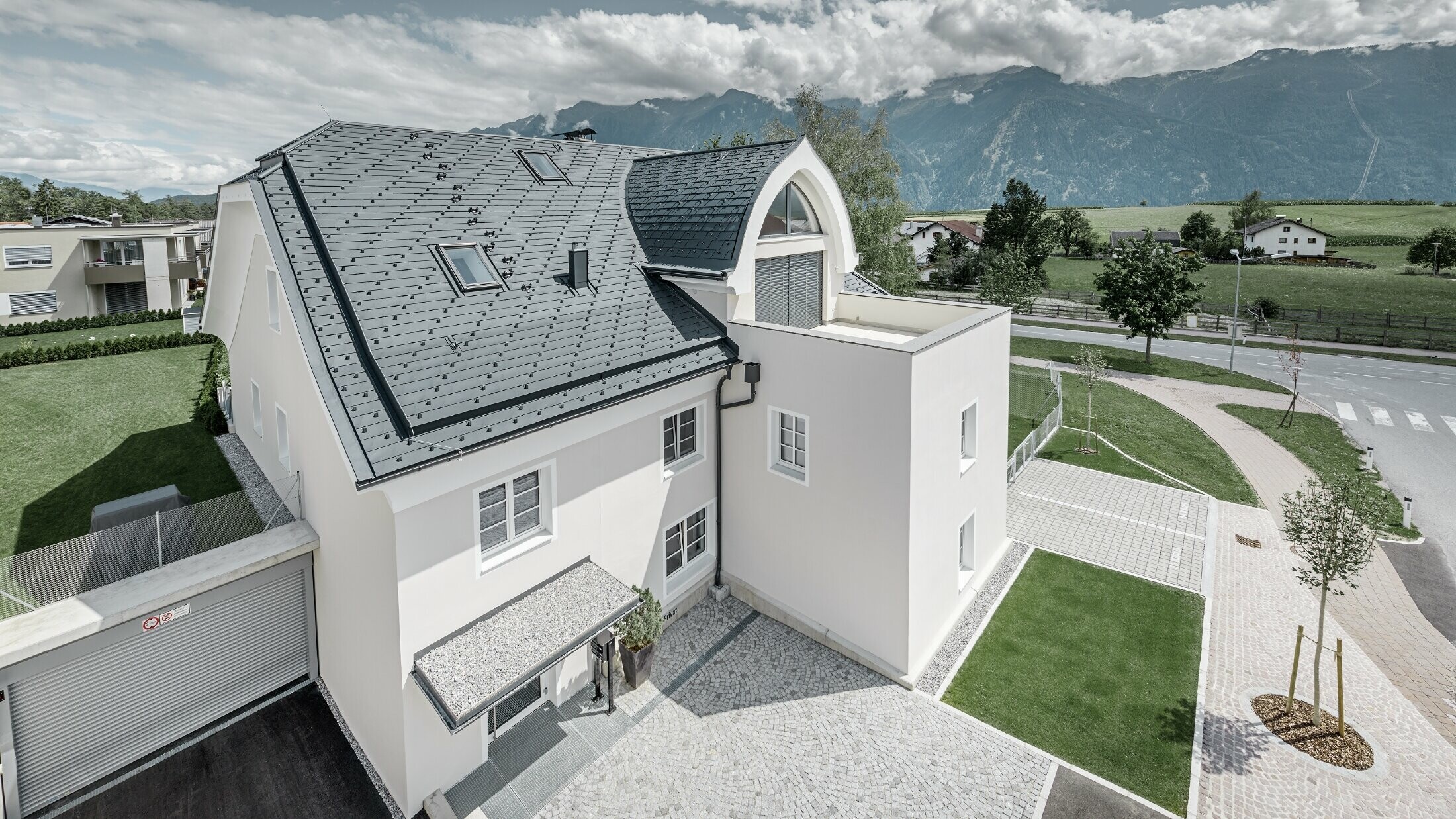 Bâtiment d’habitation rénové avec une nouvelle toiture PREFA ; le toit à demi croupe avec lucarne en ogive et loggia a été recouvert de bardeaux en alu de PREFA couleur anthracite. Des arrêts neige, des encadrements de fenêtre et des gouttières de PREFA ont également été employés.