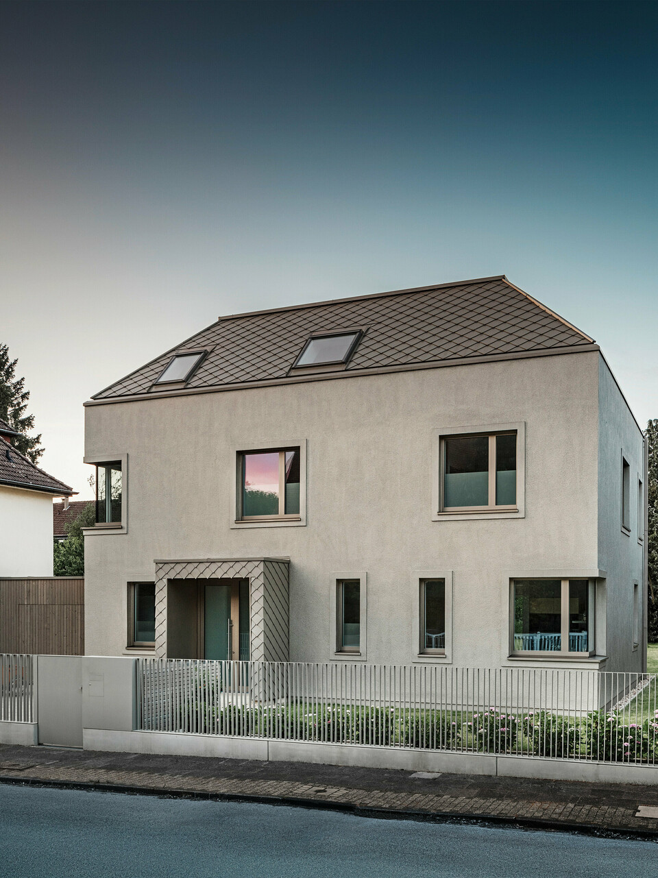 Une maison individuelle moderne avec un toit en croupe et un revêtement de toiture et de façade en aluminium couleur bronze de PREFA. La maison est située dans un quartier résidentiel calme, entourée de jardins bien entretenus et d'autres maisons individuelles. Au premier plan, vous pouvez voir une clôture basse qui sépare la propriété de la rue. L'architecture de la maison se caractérise par des lignes épurées et de grandes fenêtres qui laissent entrer beaucoup de lumière naturelle dans les intérieurs.