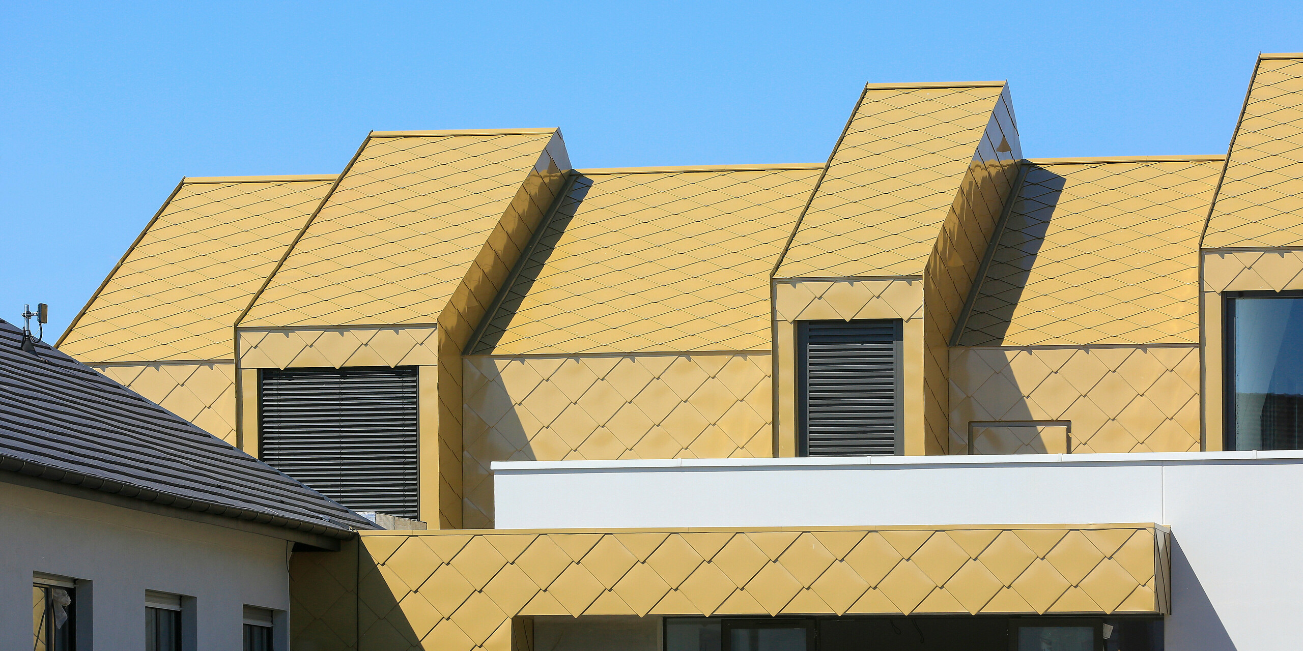 Gros plan sur le balcon et les lucarnes du Centre communautaire et culturel de Fessenheim en France. Le toit et certaines parties de la façade ont été revêtus de losanges de toiture et de façade PREFA 44 × 44 en Sparkling Gold.