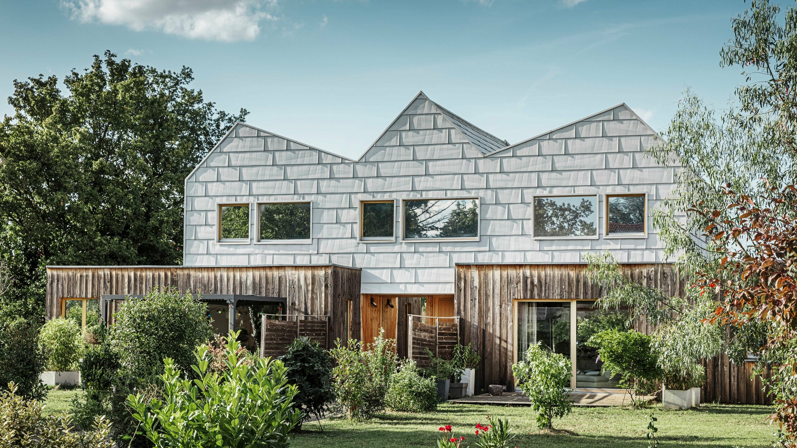 Maison jumelée moderne avec toit à pignon et façade PREFA. La façade est constituée de panneaux en aluminium FX.12, qui offrent un aspect structuré et moderne. La maison est entourée d'un jardin bien entretenu et les encadrements de fenêtres s'harmonisent parfaitement avec l'architecture globale.