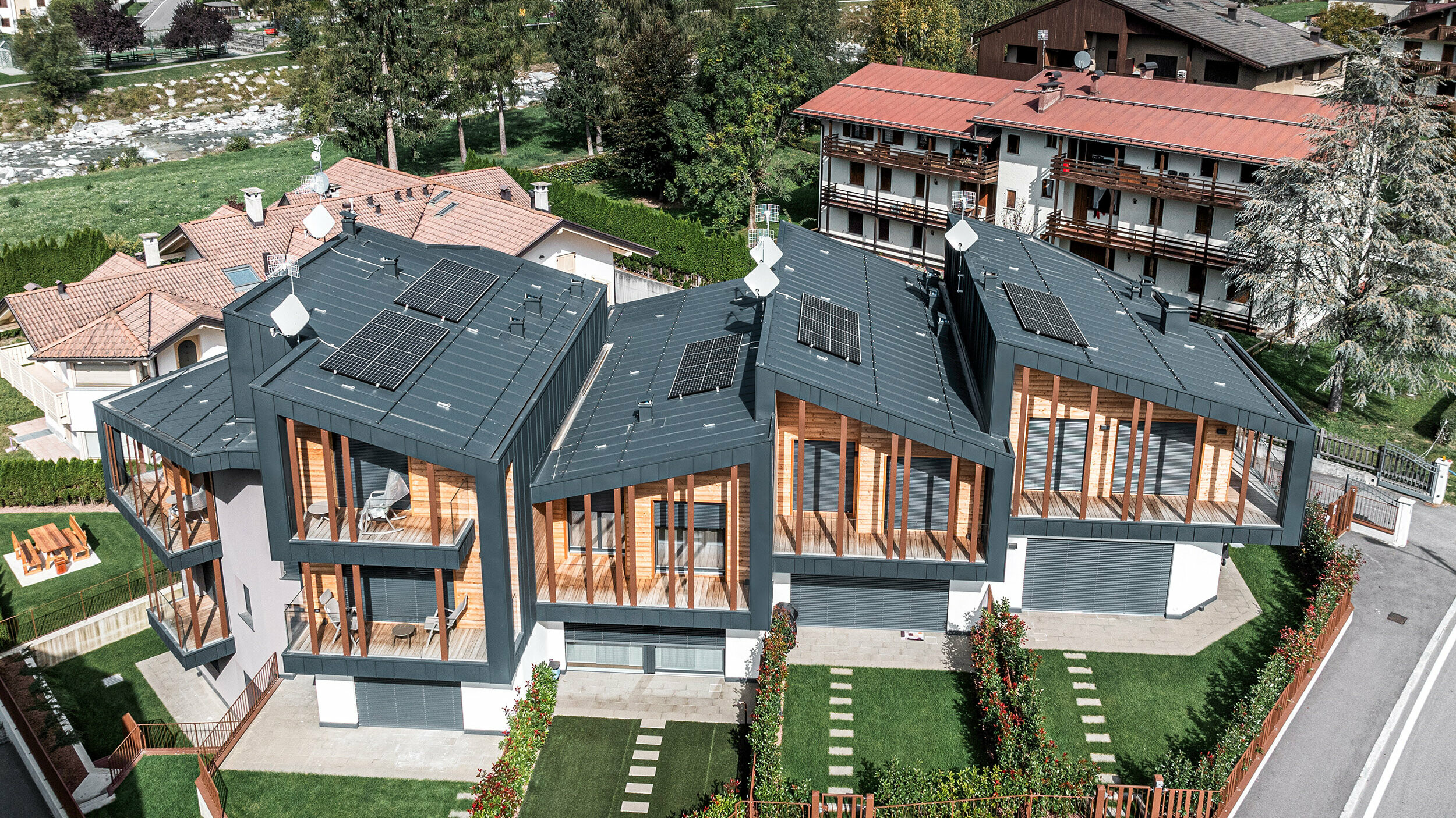 Vue aérienne de maisons de vacances modernes dans les Dolomites avec toits monopentes en métal Prefalz équipés de modules solaires. Les façades associent du métal gris foncé à des éléments naturels en bois, qui cohabitent en parfaite harmonie avec des pelouses vertes et des haies ornementales entre chaque jardin, dans un quartier en dehors du centre, caractérisé par des maisons traditionnelles et des arbres touffus en arrière-plan.