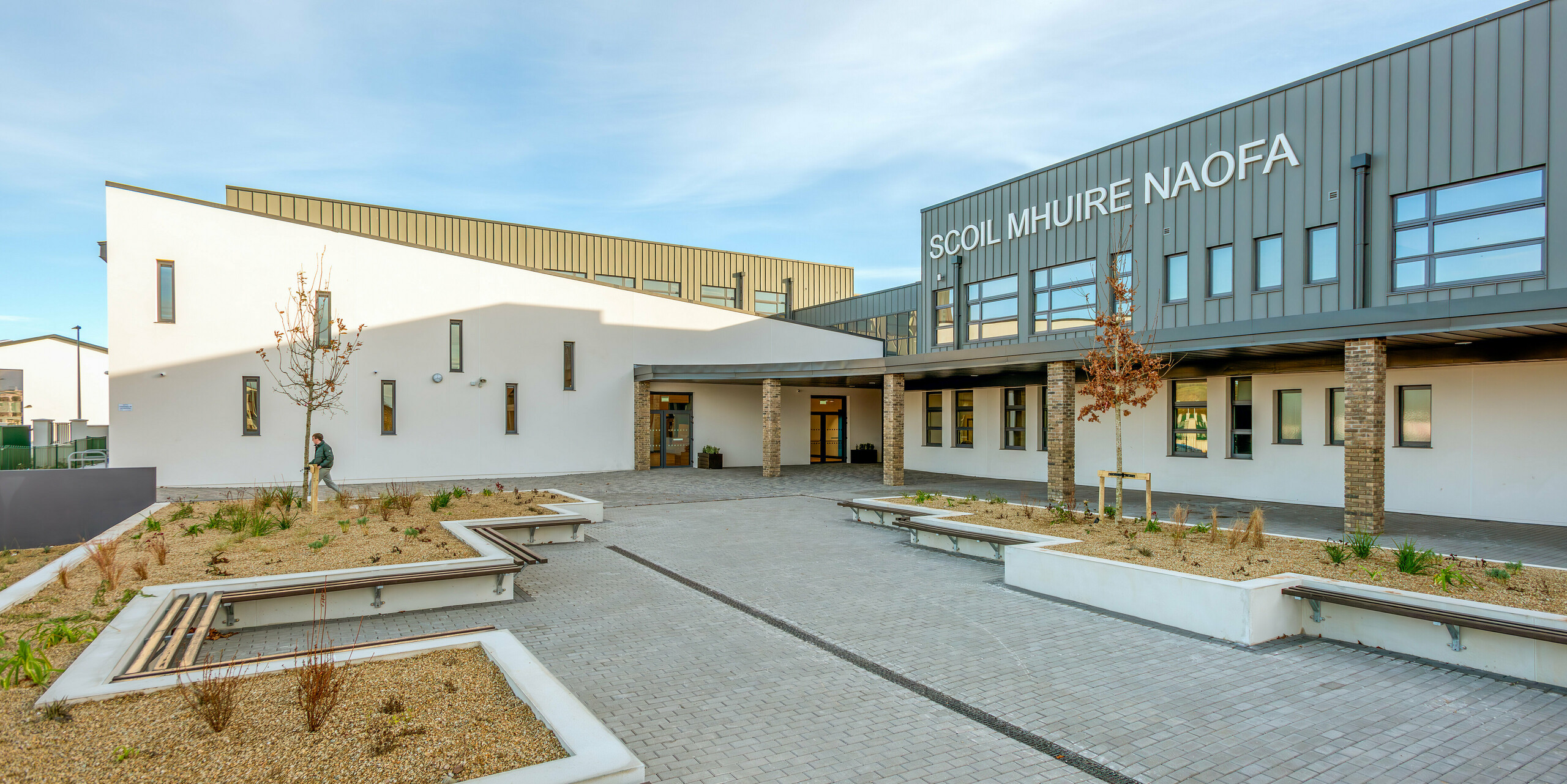 Espace extérieur accueillant de Scoil Mhuire Naofa à Carrigtwohill, en Irlande, avec des sièges et des plantations bien entretenues. La façade en aluminium PREFALZ en P.10 gris souris confère au bâtiment scolaire une élégance durable et résistante aux intempéries, complétée par des lignes claires et un design fonctionnel.