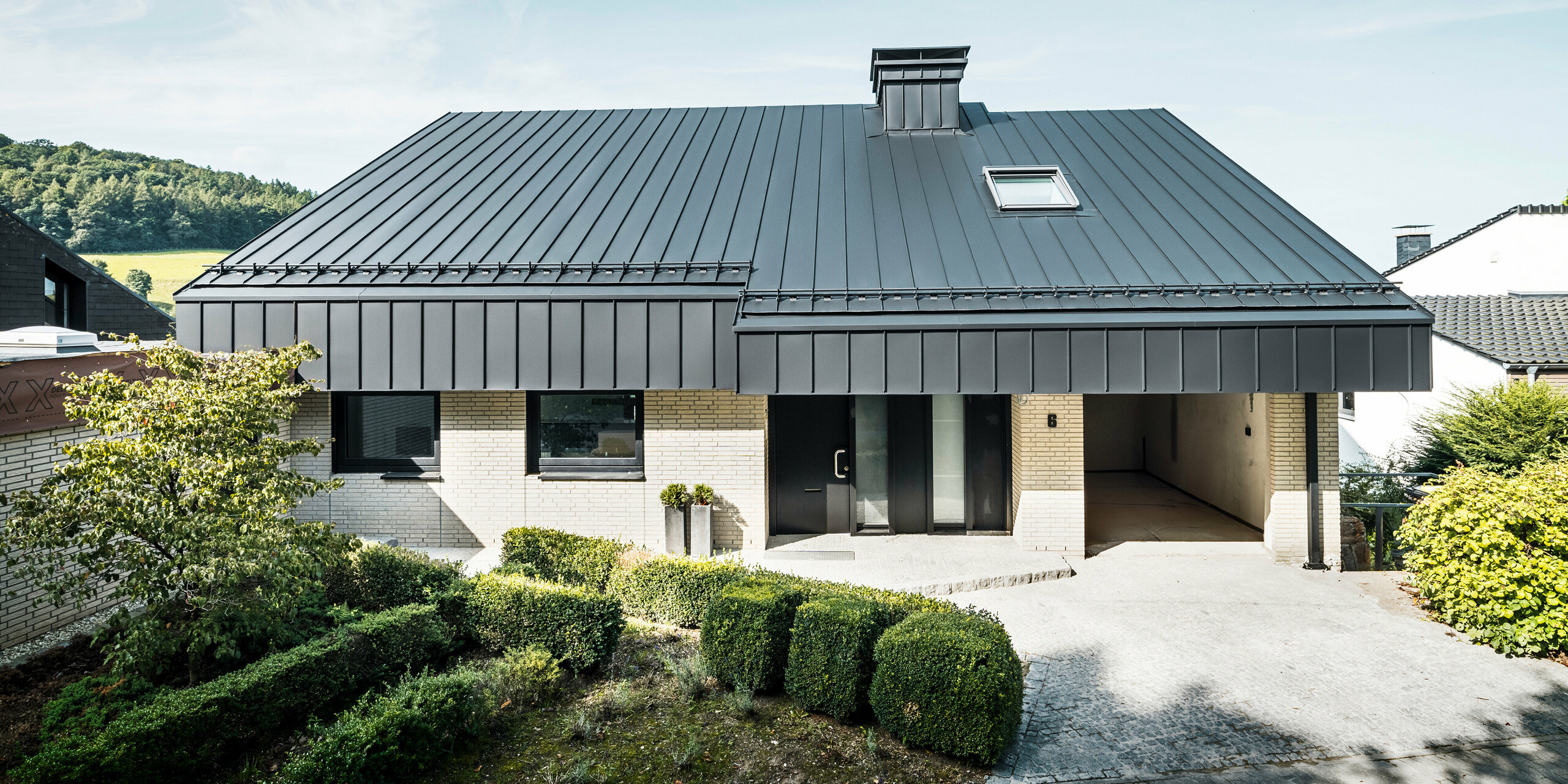 Vue de face d'une maison unifamiliale à Meschede (DE) avec toit PREFA et façade en aluminium. Pour protéger le bâtiment, le toit en appentis décalé et les deux pignons ont été recouverts d'une toiture à double joint debout PREFALZ durable dans l'élégant noir P.10. La zone d'entrée est conçue avec style avec une allée pavée et des détails architecturaux modernes. En arrière-plan, un cadre rural avec des collines vallonnées, des prairies verdoyantes et un ciel bleu vif.