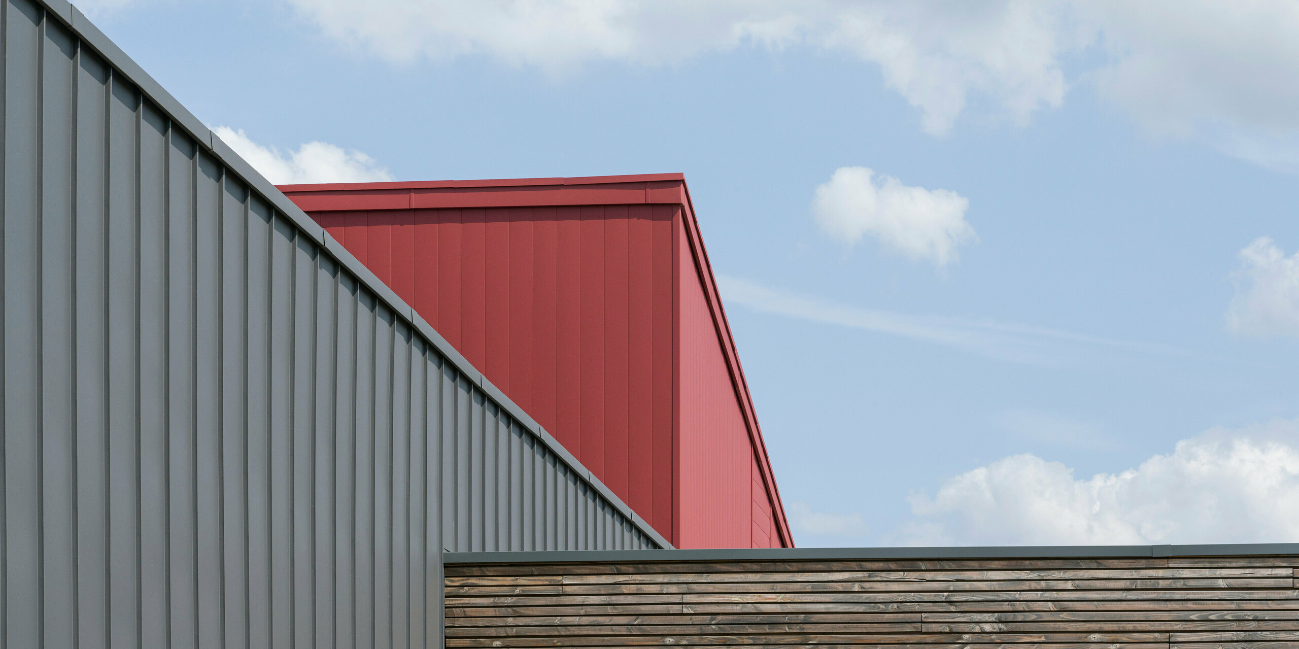 Gros plan sur la conception du toit et de la façade d'un bâtiment moderne. Au premier plan se trouve un mur avec des panneaux de bois horizontaux à la texture naturelle. Derrière elle se dresse une façade PREFALZ gris sombre aux panneaux verticaux qui respire l'élégance industrielle. Au-dessus de cette façade se trouve une partie d'un système de toiture et de façade en aluminium rouge vif, qui offre un contraste de couleurs saisissant et souligne l'architecture moderne.