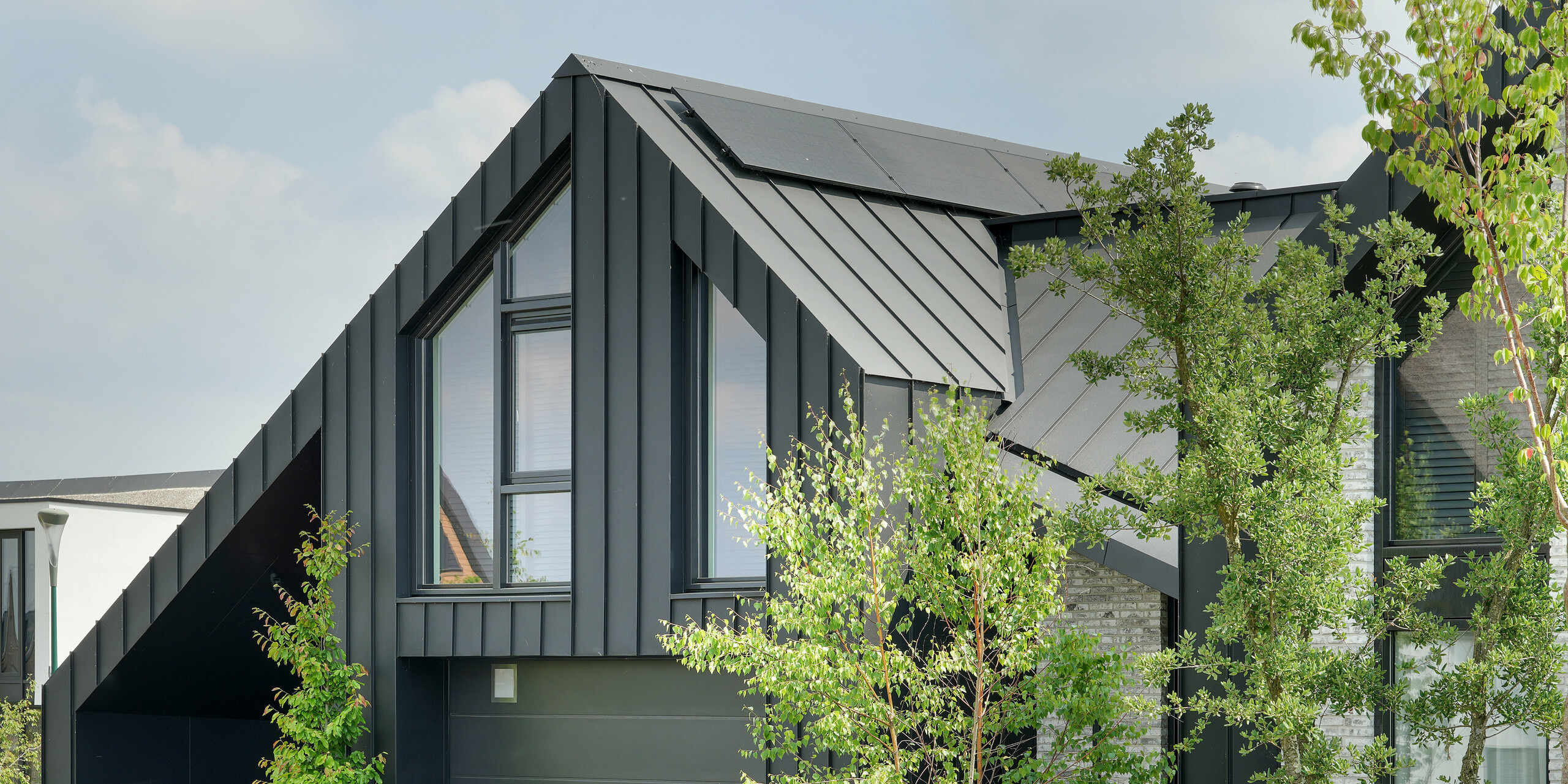 Détail d'une maison individuelle moderne à Veenendaal, Pays-Bas, présentée dans un environnement naturel. Le bâtiment se distingue par son toit pointu noir PREFALZ. Le revêtement en aluminium noir du bâtiment forme un contraste frappant avec les grandes fenêtres et la façade en pierre naturelle gris clair. Le mélange varié de surfaces sombres et claires crée une juxtaposition captivante, encore accentuée par l'espace vert environnant.