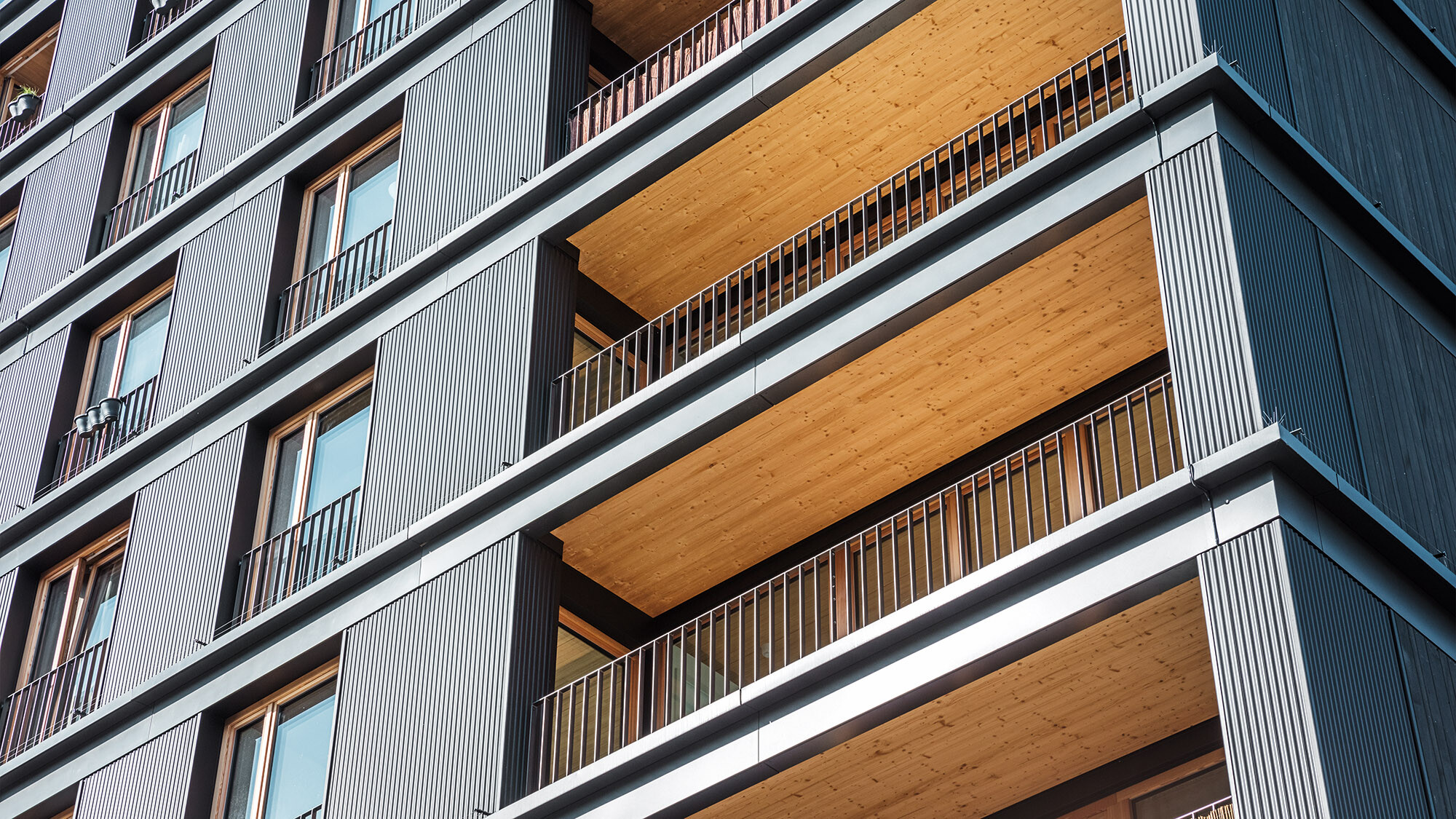 Le bâtiment multifonctionnel vu de côté en perspective de grenouille : le profil ondulé, les balcons et les plafonds de balcons revêtus de bois sont en vue.