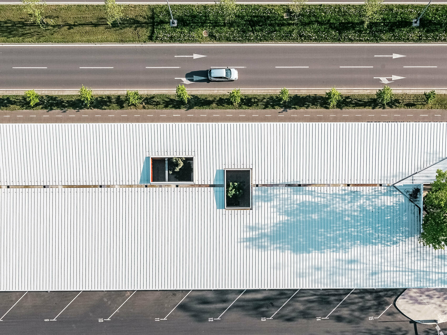 La photo montre une vue aérienne du hall de la gare routière en Croatie recouvert d'un toit Prefalz blanc de PREFA. Le long bâtiment intègre plusieurs ouvertures rectangulaires disposées de manière asymétrique, laissant transparaître le feuillage vert des arbres. On distingue un parking plus petit au premier plan, tandis qu'une route à deux voies, bordée d'une rangée d'arbres, longe le hall à l'arrière-plan.