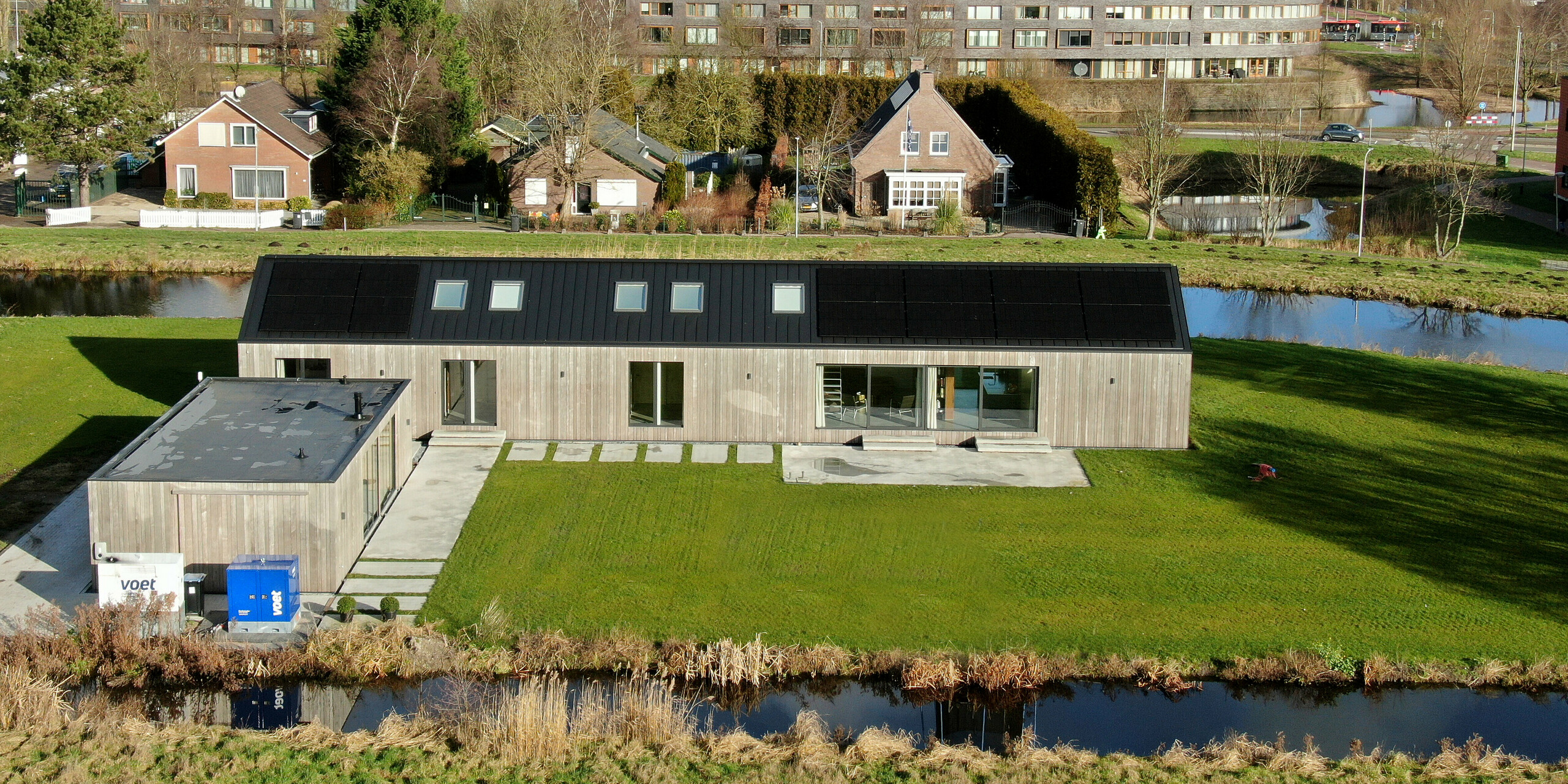 Vue aérienne d'un bungalow moderne comprenant une dépendance à Uithoorn, aux Pays-Bas, avec toiture à joints debout en PREFALZ gris noir et système photovoltaïque intégré. La photo montre l'ensemble du complexe avec un grand jardin et des douves environnantes. La maison se caractérise par de grandes façades vitrées et une façade en bois naturel, qui créent une connexion harmonieuse avec l'environnement. Idéal pour les projets de construction durable intégrant les énergies renouvelables et un design moderne.