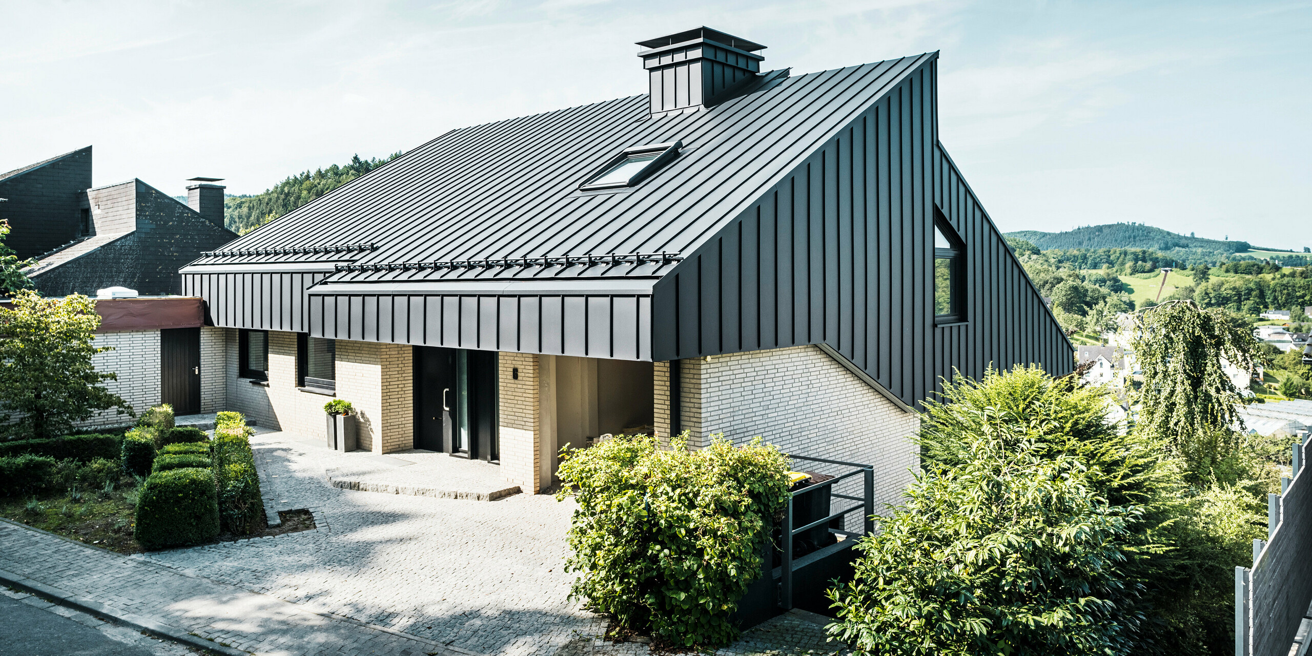 Architecture moderne d'une maison unifamiliale à Meschede, en Allemagne, avec un élégant toit en pente décalé en PREFALZ P.10 noir. La façade en aluminium s'harmonise avec les lignes épurées du bâtiment, tandis que les environs avec des haies vertes et des plantes complètent le tableau général.