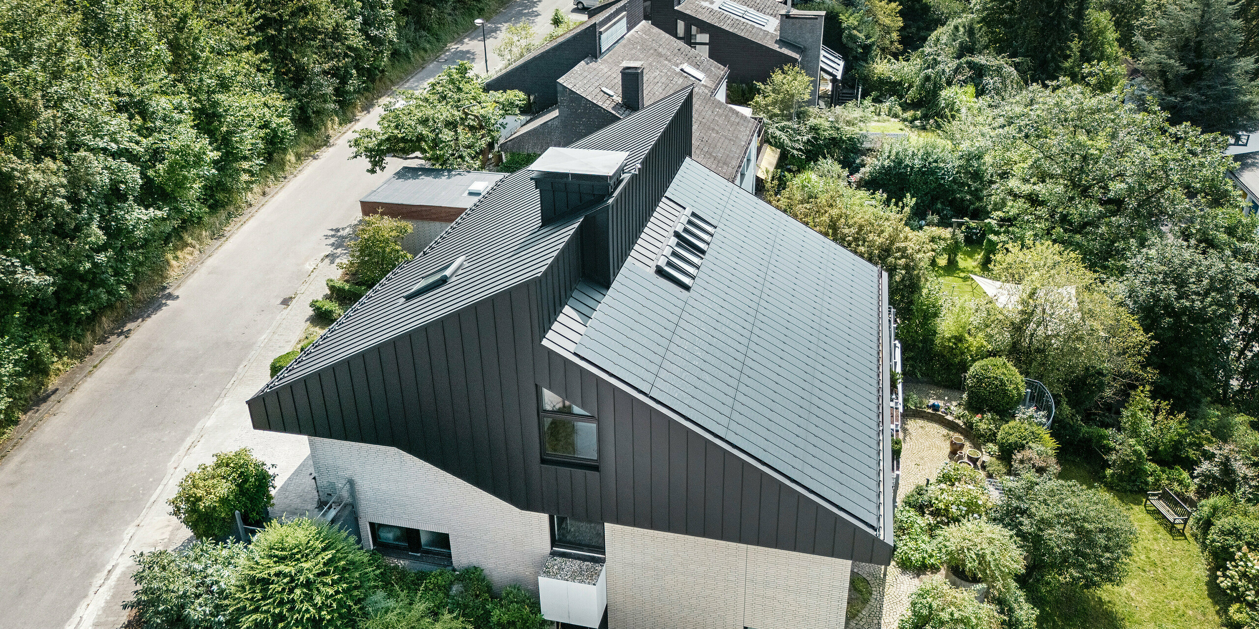 Vue oblique d'une maison unifamiliale moderne à Meschede, en Allemagne. Le toit en pente décalé a été revêtu de PREFALZ en P.10 noir. Le toit en aluminium constitue un élément visuel marquant et offre au bâtiment une protection permanente. La toiture à double joint debout de précision s'étend du toit à travers la façade jusqu'aux pignons. Entouré d'une végétation luxuriante et d'une rue calme, le bâtiment économe en énergie avec les modules solaires innovants PREFALZ s'intègre parfaitement dans l'environnement de vie naturel.