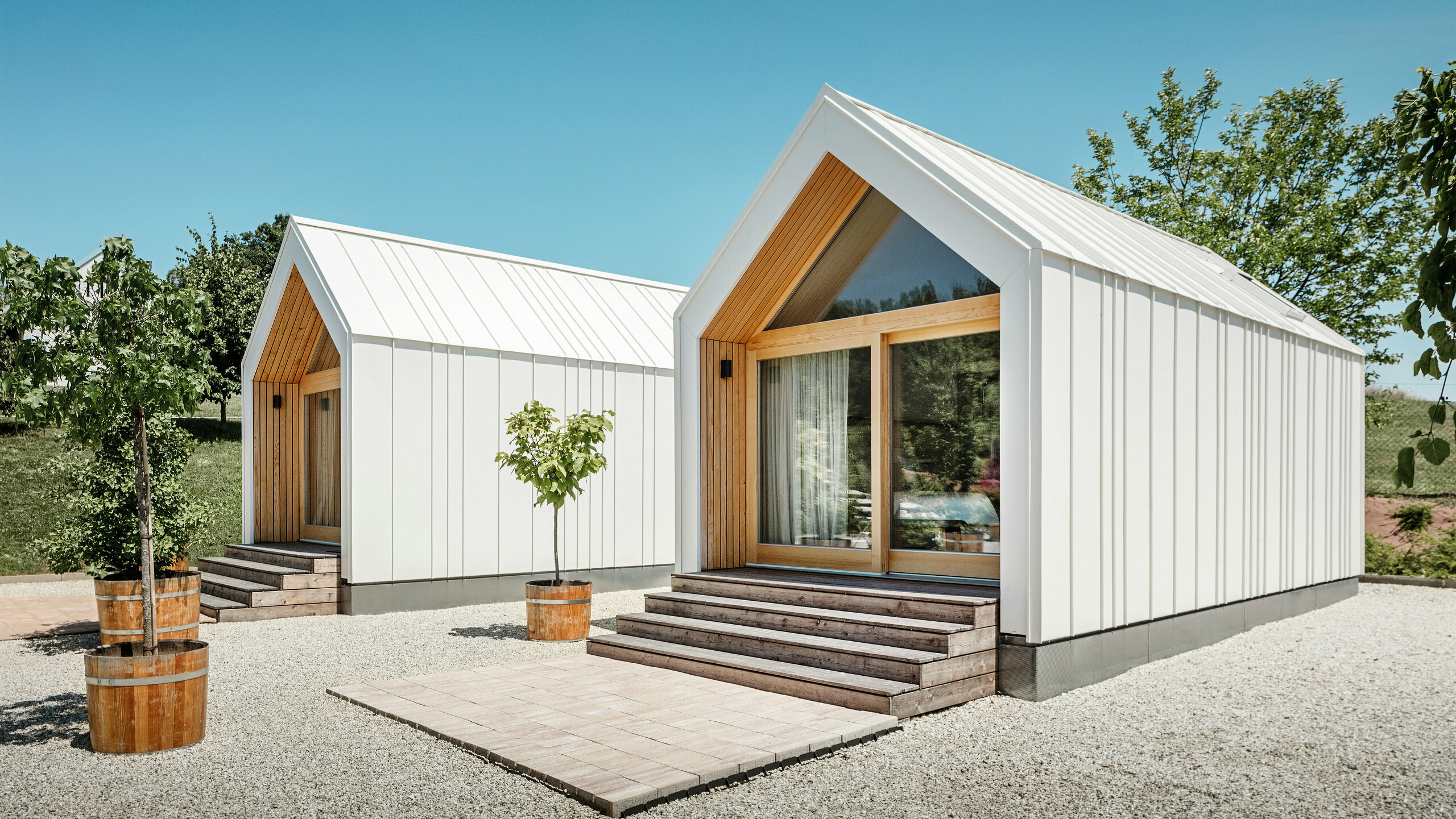 Deux petites maisons blanches dans l'idylle de vacances « Pri Momi » à Kančevci, en Slovénie, conçues avec un revêtement de toit et de façade PREFALZ en blanc pur P.10 et en bois naturel. L'architecture minimaliste des bâtiments est soulignée par l'élégant mélange de matériaux aluminium et bois. Les grandes façades vitrées aux entrées ainsi que les marches en bois et les arbres environnants créent une atmosphère invitante et harmonieuse. L'hébergement exclusif est situé au milieu d'un paysage pittoresque et est utilisé spécifiquement pour les célébrations.