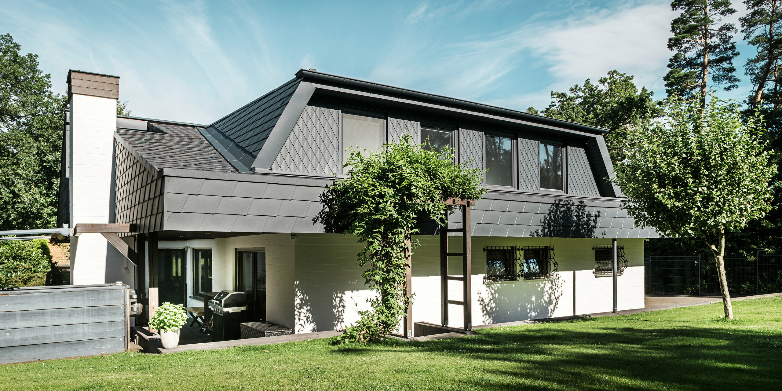 Vue latérale d'une maison individuelle à Mechernich avec des bardeaux DS.19 PREFA et de petits losanges de toiture en anthracite P.10. Les lucarnes trapézoïdales sont recouvertes de losanges de toit en tôle robuste, tandis que le toit est recouvert de bardeaux. Une pergola verte et des arbres dans le jardin complètent l'architecture moderne et résistante aux intempéries.