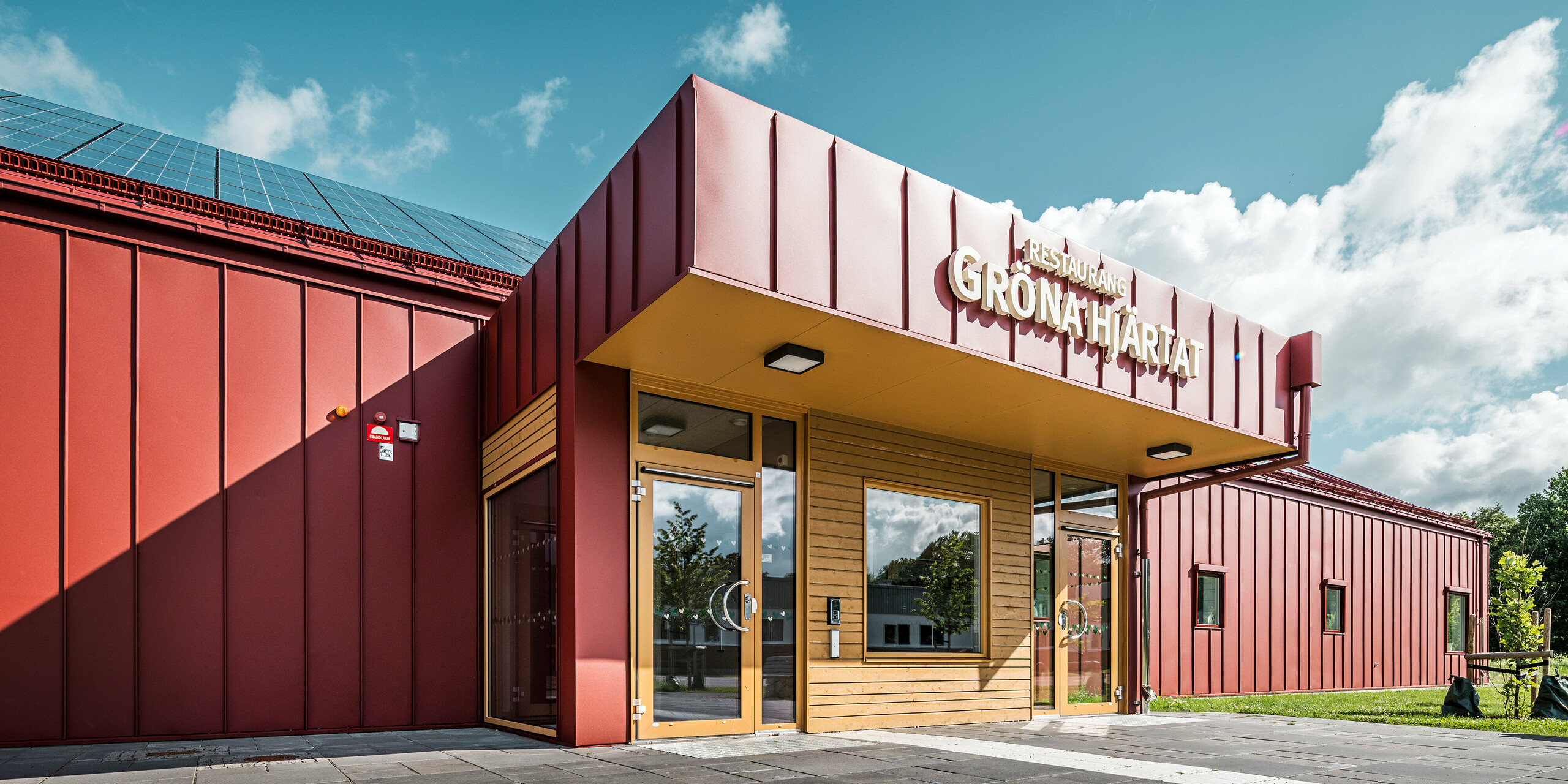 Gros plan de la zone d'entrée du restaurant Gröna Hjärtat, de la cantine scolaire de Göingeskolan et Prästavångskolan à Broby, en Suède. La façade et le toit sont revêtus de PREFALZ en rouge oxyde, ce qui confère au bâtiment un aspect moderne et marquant. Le panneau « Restaurang Gröna Hjärtat » est apposé au-dessus de l'entrée. La combinaison d'un revêtement en aluminium durable et d'une conception économe en énergie, y compris un système photovoltaïque installé sur le toit, fait du bâtiment un exemple exceptionnel d'architecture contemporaine. a gouttière PREFA est conçu en rouge oxyde et contribue à l'aspect général harmonieux.