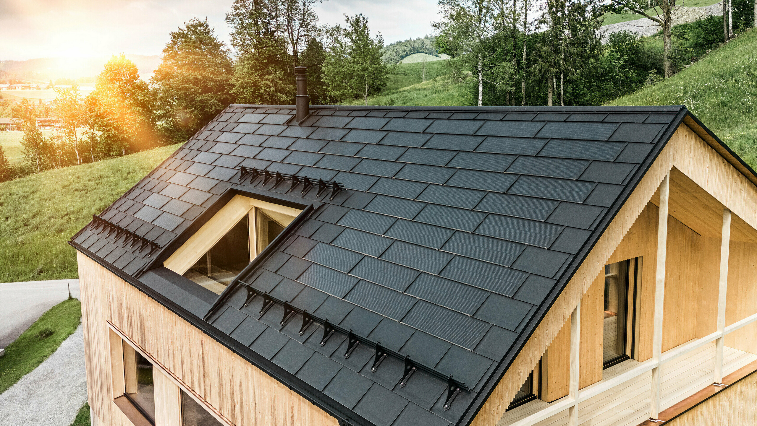 Prise de vue d'une maison en bois moderne et durable à Egg avec un toit solaire PREFA, qui se distingue par son aspect sombre, structuré et élégant. Les modules solaires sont stratégiquement placés entre les R.16 classiques pour profiter d'un rayonnement solaire optimal et s'intégrer esthétiquement dans la conception globale. La maison est en harmonie avec son environnement verdoyant, tandis que le chaud soleil du soir met en valeur la texture naturelle du bois de la façade et souligne la solidité de la construction. Un exemple harmonieux de bâtiment respectueux de l’environnement et d’efficacité énergétique.