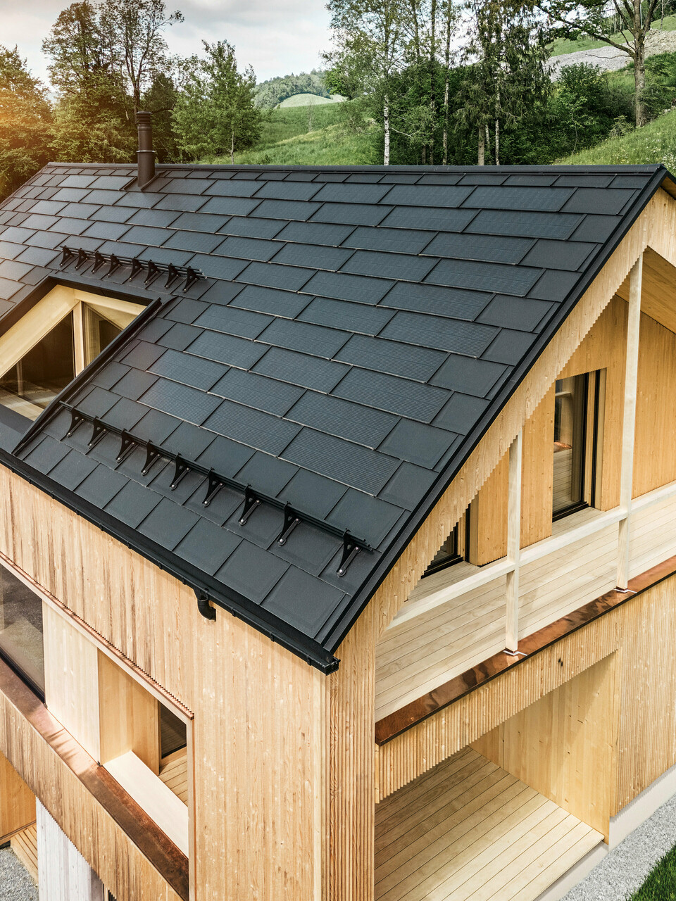 Prise de vue d'une maison en bois moderne et durable à Egg avec un toit solaire PREFA, qui se distingue par son aspect sombre, structuré et élégant. Les modules solaires sont stratégiquement placés entre les R.16 classiques pour profiter d'un rayonnement solaire optimal et s'intégrer esthétiquement dans la conception globale. La maison est en harmonie avec son environnement verdoyant, tandis que le chaud soleil du soir met en valeur la texture naturelle du bois de la façade et souligne la solidité de la construction. Un exemple harmonieux de bâtiment respectueux de l’environnement et d’efficacité énergétique.