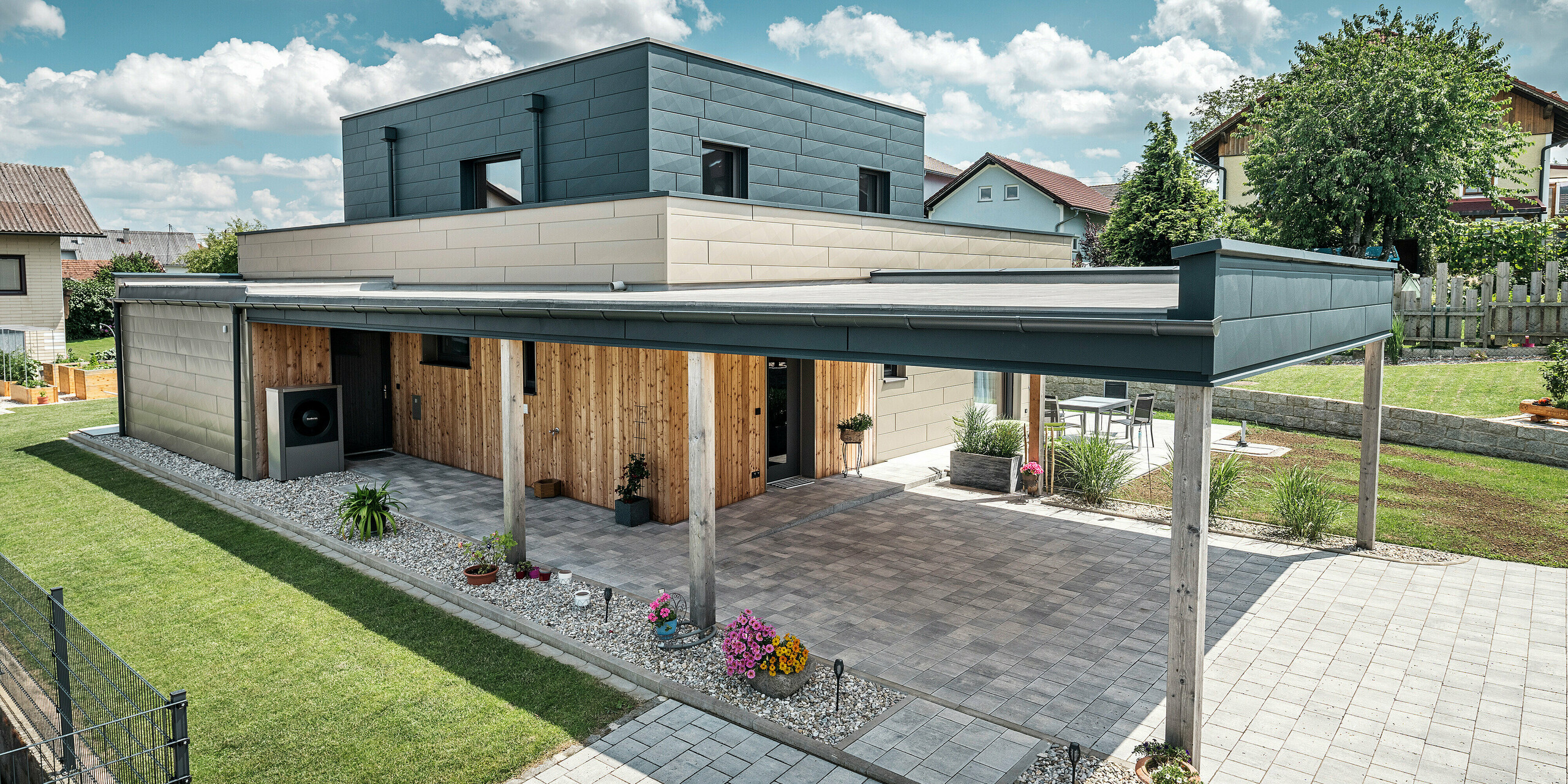 Vue aérienne de l'allée d'une maison familiale moderne à Wippenham, en Autriche, avec une façade en aluminium PREFA bicolore bronze et anthracite. La maison montre l'intégration élégante des panneaux de façade Siding.X et du revêtement en bois. Le carport couvert, parfaitement intégré à l'architecture de la maison, est clairement visible. Le jardin et la terrasse bien entretenus soulignent le design moderne et fonctionnel du bâtiment.