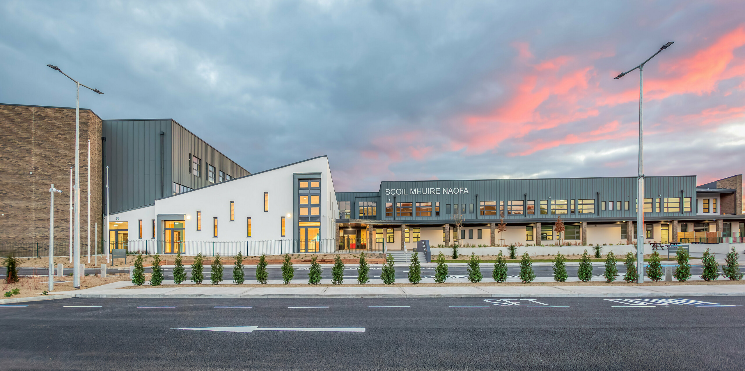 Vue de face du nouveau Scoil Mhuire Naofa à Carrigtwohill, en Irlande, au coucher du soleil. La façade en aluminium PREFALZ gris souris associée à des détails de brique modernes et à des lignes claires crée une architecture harmonieuse et durable. L’espace extérieur spacieux souligne l’atmosphère accueillante de l’école.