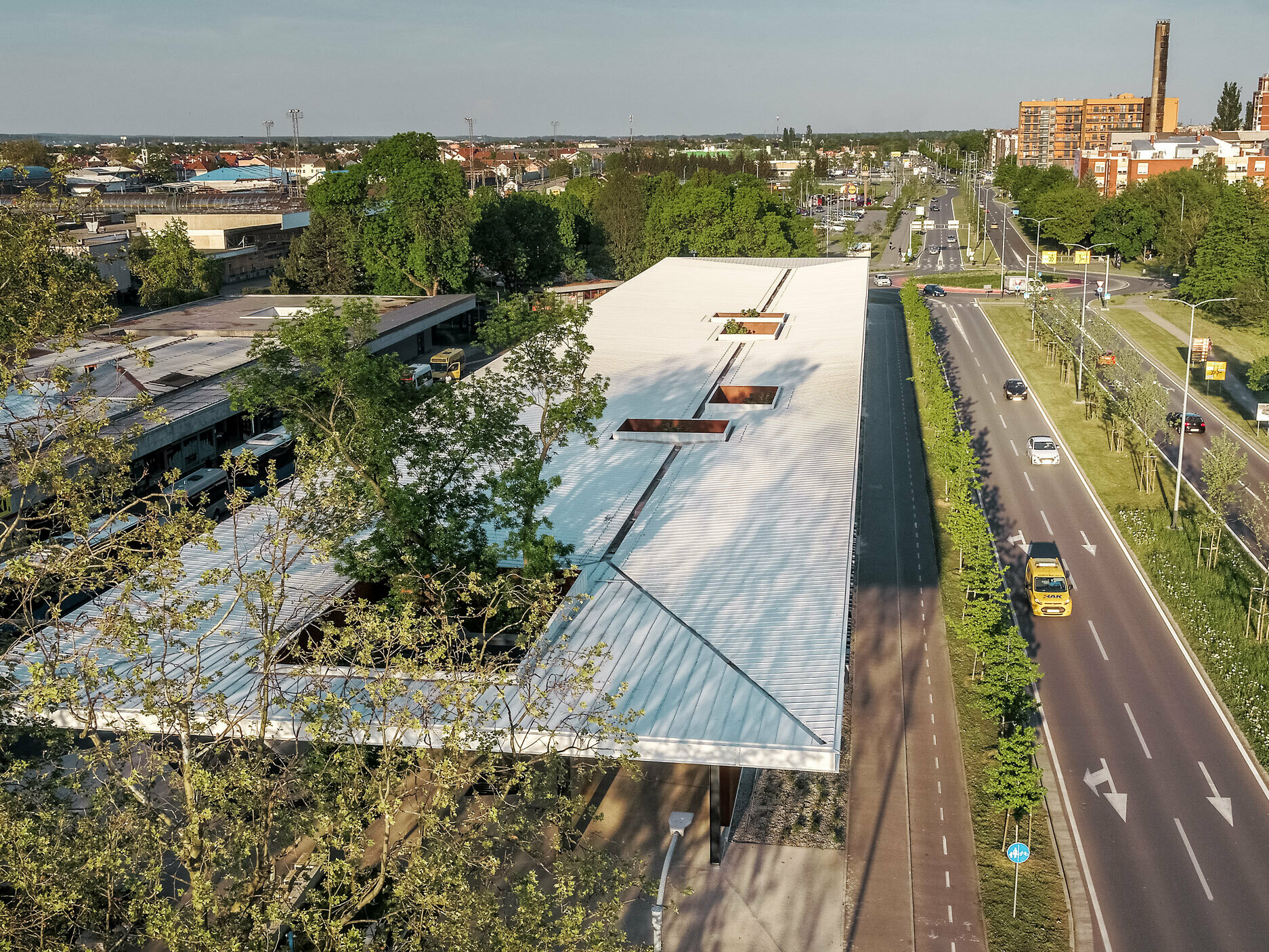 La photo offre une vue panoramique d'un hall de gare routière moderne en Croatie, recouvert d'un toit Prefalz blanc de PREFA. Le toit est percé de plusieurs ouvertures rectangulaires d'où jaillissent des arbres. Le hall de gare jouxte une large route à deux voies bordée d'arbres. À l'arrière-plan, on peut voir divers bâtiments et habitations résidentielles caractéristiques de l'environnement urbain.