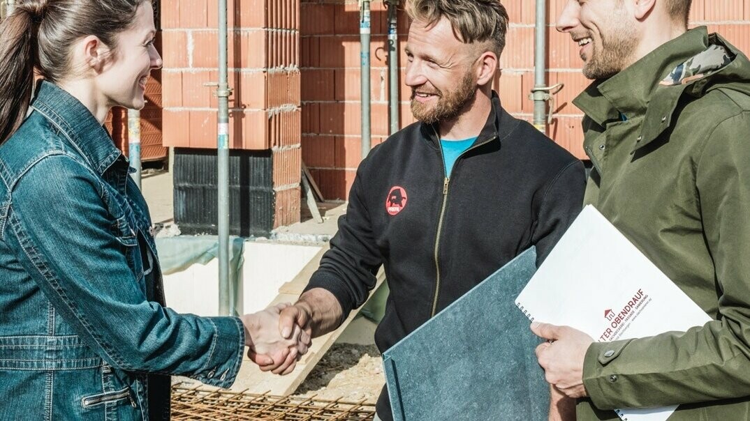 Ein junges Pärchen trifft sich auf ihrer Hausbau-Baustelle mit dem Handwerker in der Nähe. Sie haben das Angebot besprochen und angenommen. Der Spengler hält eine PREFA Dachplatte in der Hand.