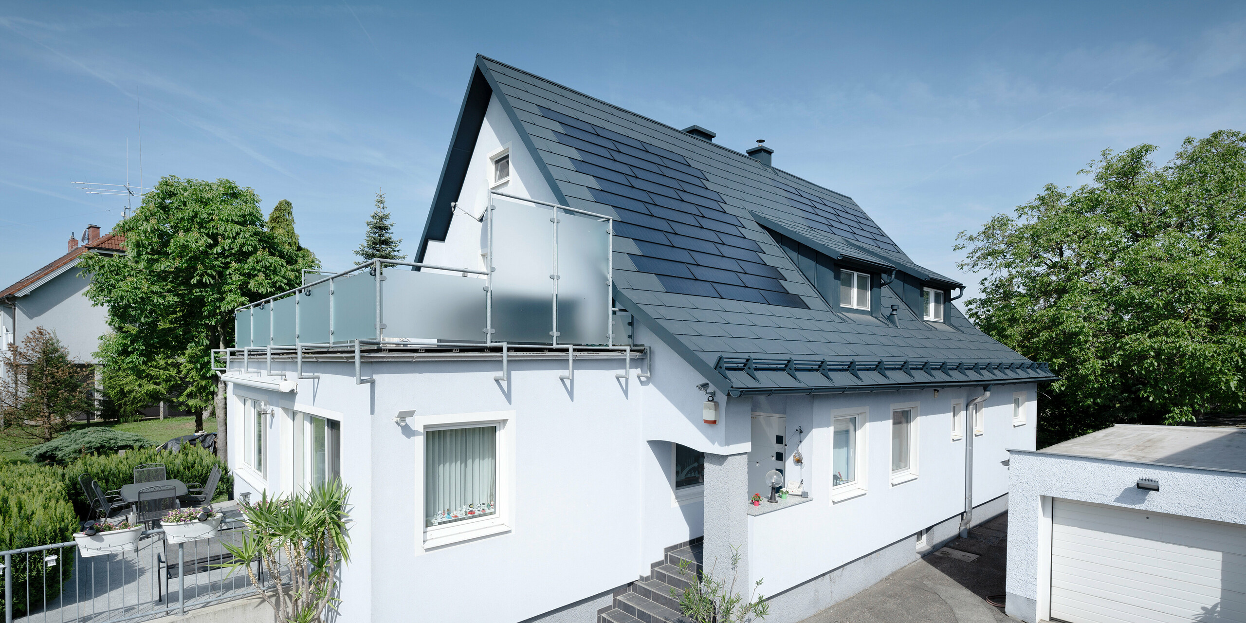 Rénovation du toit d'une maison individuelle à Leonding avec des tuiles solaires PREFA en anthracite. La photo montre la maison rénovée d'un point de vue latéral avec une vue dégagée sur le toit et les modules solaires intégrés. Ceux-ci offrent un moyen durable et économe en énergie de produire de l’électricité. Dans cette propriété, les tuiles solaires génèrent une puissance électrique de 10,6 kWc. Les matériaux PREFA modernes, durables et résistants aux intempéries contribuent à une construction de toiture esthétique et fonctionnelle qui répond aux dernières normes en matière de construction durable.