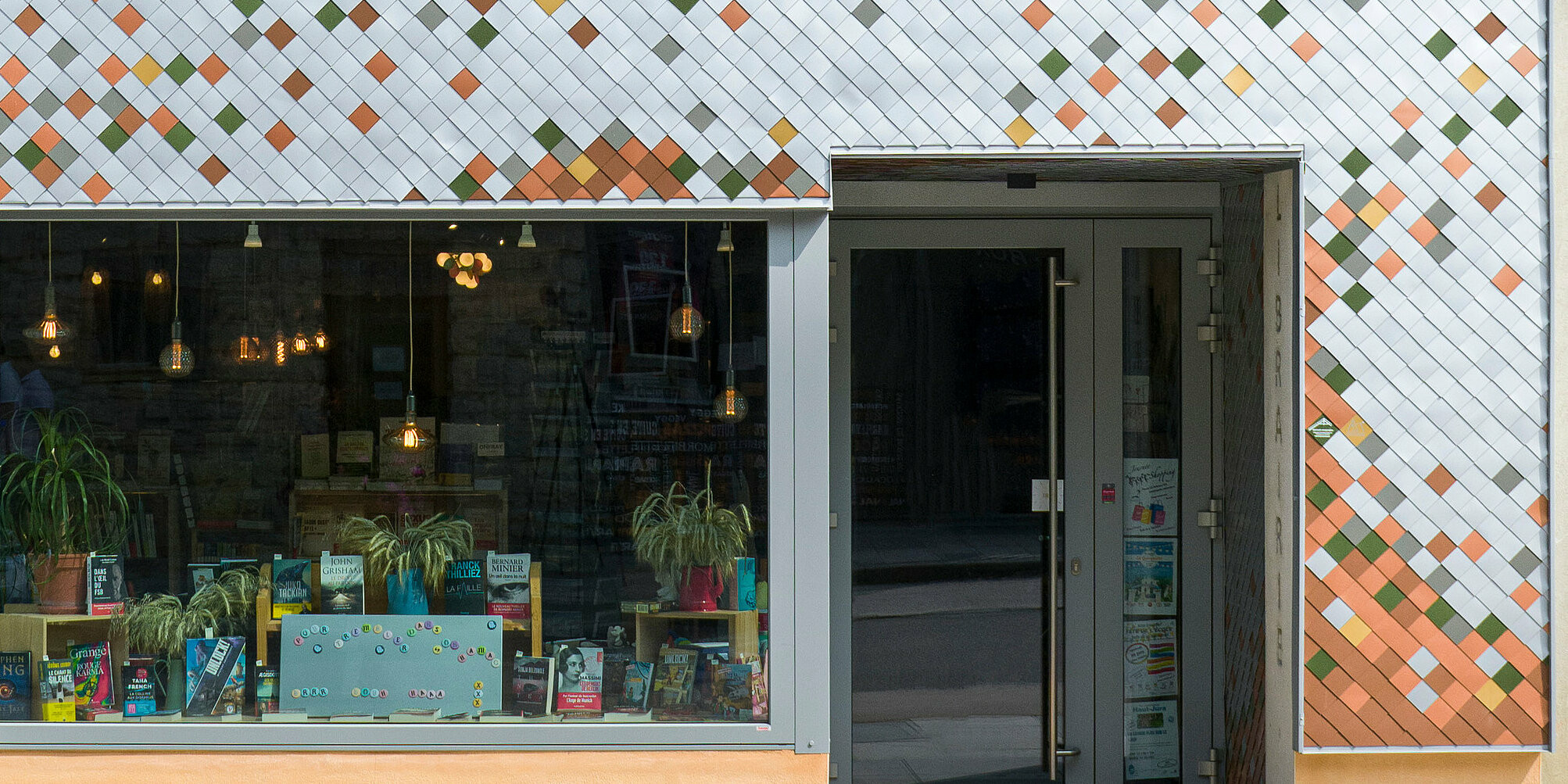 Vue de face de la façade colorée d'une librairie de la petite communauté française des Rousses. Le bâtiment combine un étage supérieur orange vif avec des éléments décoratifs et un étage inférieur avec une façade en forme de mosaïque en aluminium de différentes couleurs. Une grande fenêtre au rez-de-chaussée présente des livres et des plantes à côté de la porte d'entrée. Une étroite gouttière longe le côté du bâtiment et le numéro 55 est visible sur un panneau vert à côté de la porte.