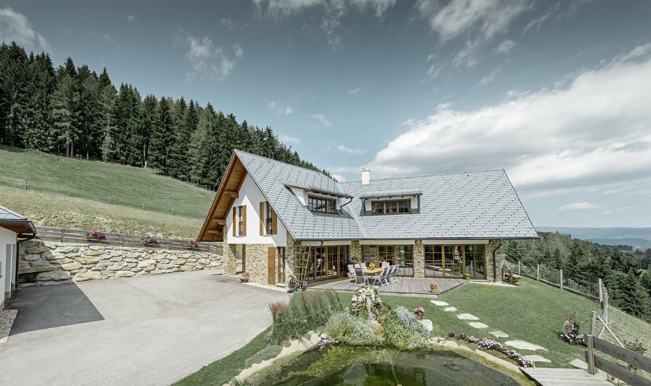 Construite en forme de L et à flanc de coteau, cette maison individuelle présente un toit à deux pans avec deux lucarnes rampantes. La toiture a été recouverte de bardeaux de toiture PREFA couleur gris pierre. Un étang se trouve au premier plan et on peut voir une forêt en arrière plan.