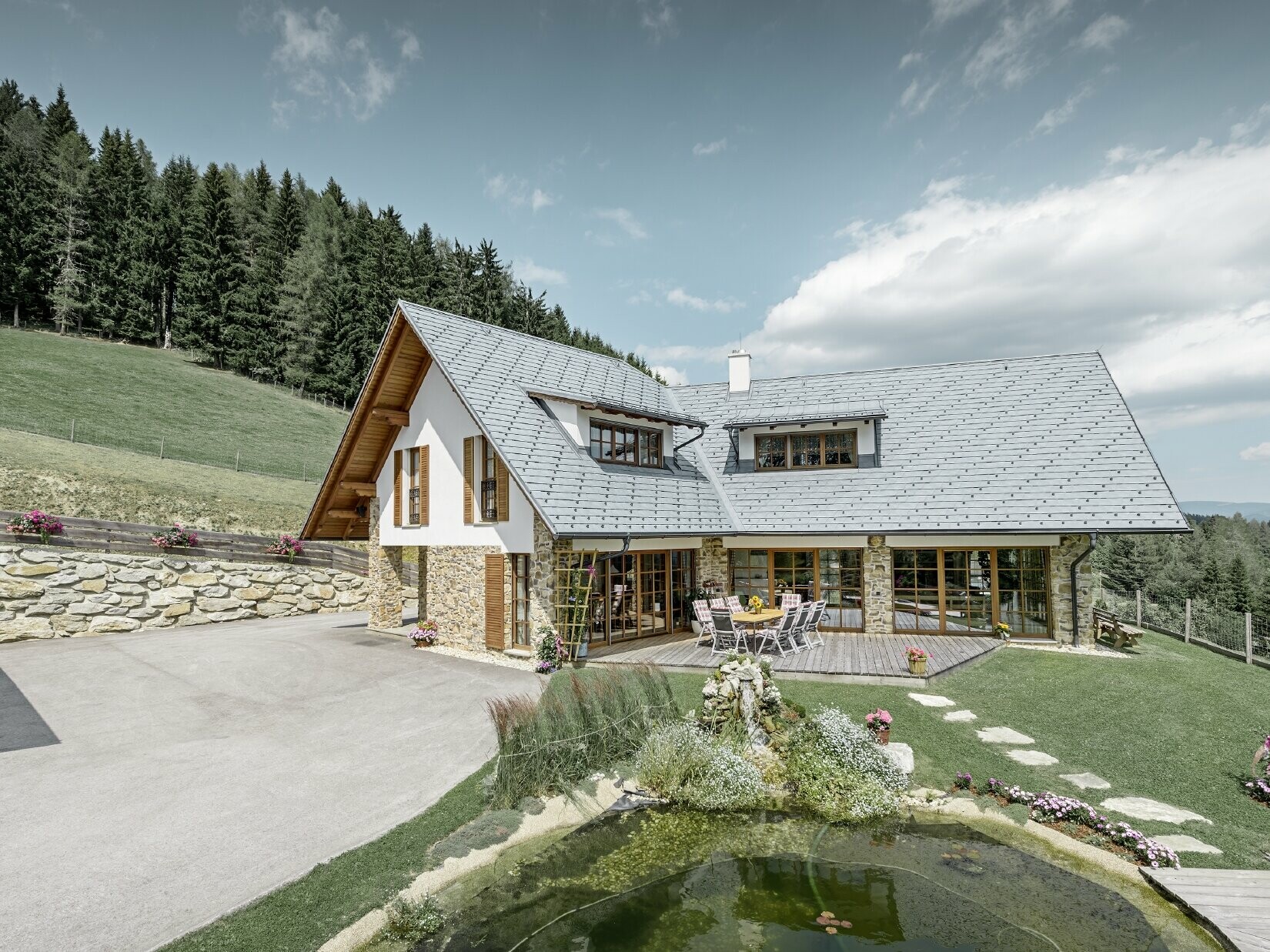 Construite en forme de L et à flanc de coteau, cette maison individuelle présente un toit à deux pans avec deux lucarnes rampantes. La toiture a été recouverte de bardeaux de toiture PREFA couleur gris pierre. Un étang se trouve au premier plan et on peut voir une forêt en arrière plan.