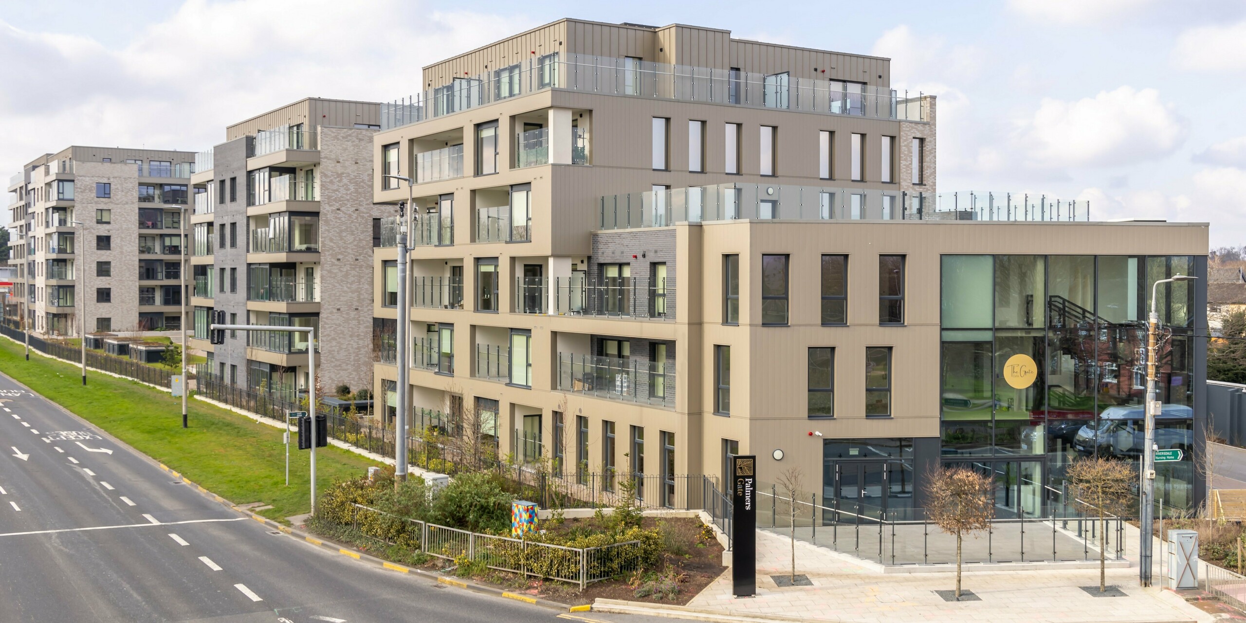 Vue extérieure du complexe résidentiel Palmers Gate à Palmerstown, Dublin, Irlande. Le complexe comprend plusieurs bâtiments résidentiels revêtus de revêtements PREFA de couleur bronze et de systèmes de façade PREFALZ. Les façades combinent un revêtement en aluminium avec des éléments en brique et de grandes baies vitrées qui soulignent le style moderne et élégant. Les immeubles offrent des balcons avec balustrades en verre et des espaces communs spacieux. La zone d'entrée du complexe présente de grandes façades vitrées et un panneau portant l'inscription « Palmers Gate ». Au total, environ 1 700 m² de revêtement PREFA et environ 1 200 m² de système de façade PREFALZ ont été utilisés.