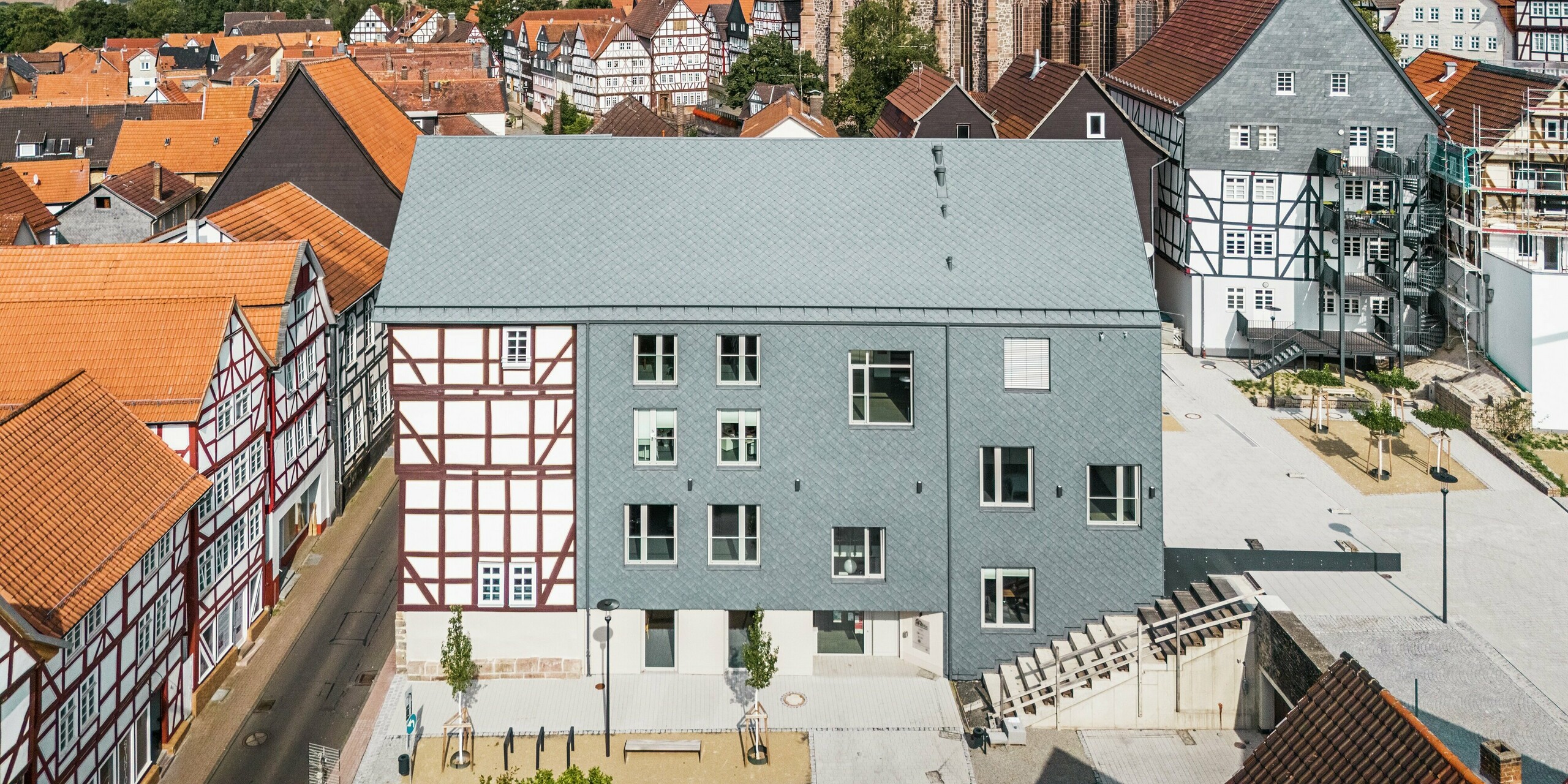 [Translate to Français:] Drohnenaufnahme des Volkshauses in Homberg, DE, mit PREFA Dach- und Wandrauten 29 × 29 in P.10 Steingrau. Das modern sanierte Gebäude harmoniert dank der langlebigen und witterungsbeständigen Aluminiumbekleidung perfekt mit den umliegenden Fachwerkhäusern und der historischen Kirche.