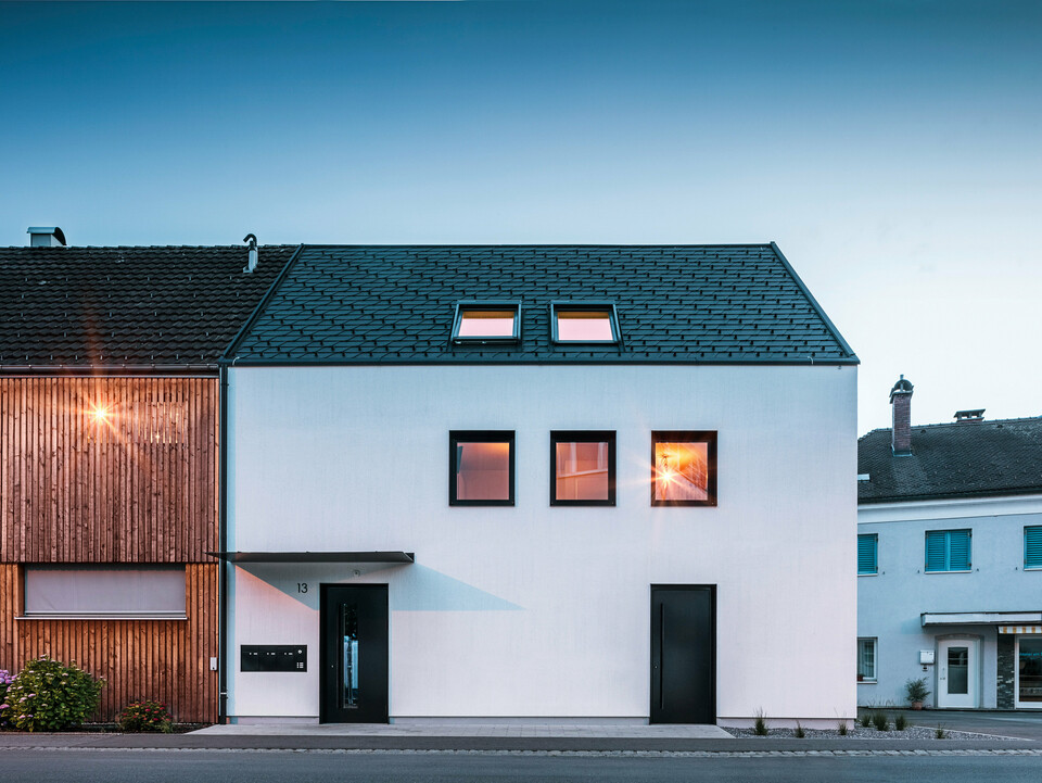 Façade moderne d'un immeuble d'habitation avec bardeaux PREFA DS19 et Prefalz en noir au bord du lac de Constance en Autriche. Le bâtiment est clairement divisé en deux, avec une extension en bois sur la gauche et une partie principale crépie sur la droite. Le toit présente la couverture en bardeaux foncés caractéristique et il y a plusieurs fenêtres rectangulaires qui se reflètent dans la chaude lumière du soir.