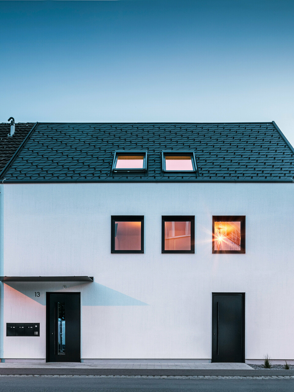 Façade moderne d'un immeuble d'habitation avec bardeaux PREFA DS19 et Prefalz en noir au bord du lac de Constance en Autriche. Le bâtiment est clairement divisé en deux, avec une extension en bois sur la gauche et une partie principale crépie sur la droite. Le toit présente la couverture en bardeaux foncés caractéristique et il y a plusieurs fenêtres rectangulaires qui se reflètent dans la chaude lumière du soir.