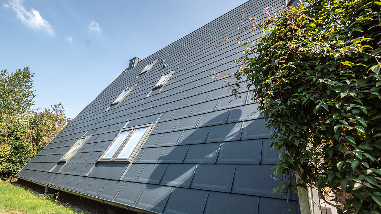 Longue surface de toit d’une maison à ossature en A recouverte de tuiles PREFA couleur P.10 anthracite. Quelques fenêtres de toit sont intégrées dans la surface de toit.