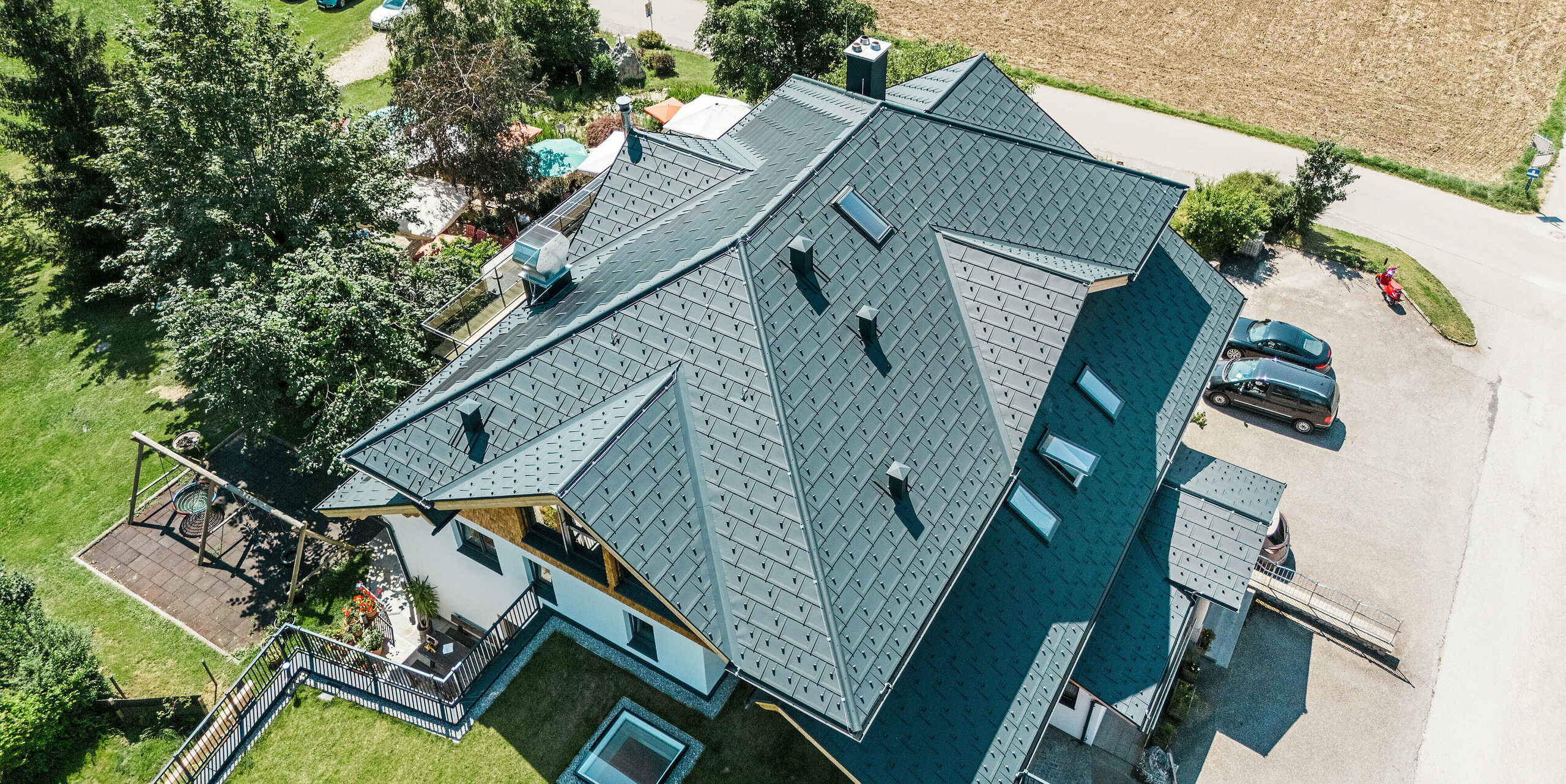 Vue d'en haut du toit du Zenz'n Stub'n à Schörfling am Attersee, recouvert de panneaux de toiture PREFA R.16 et de tuiles solaires avec revêtement de haute qualité P.10 anthracite. Le toit innovant en aluminium combine une esthétique moderne avec une technologie solaire durable et s'intègre harmonieusement dans l'environnement rural. Le système de toiture durable PREFA offre une protection complète et une apparence élégante.