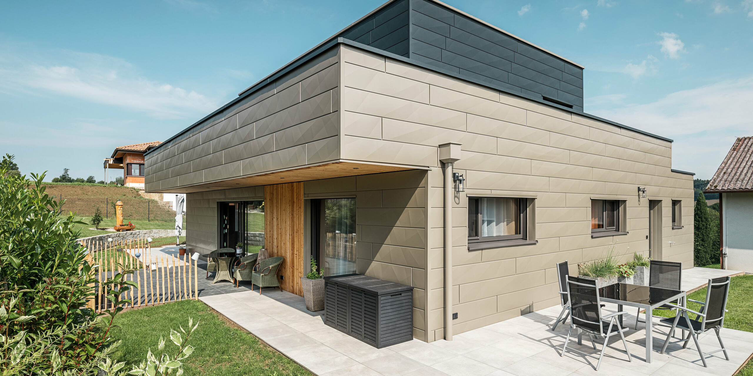 Une maison individuelle moderne à Wippenham, en Autriche, avec une façade en aluminium PREFA bicolore, composée des panneaux de façade Siding.X en bronze et anthracite. La façade présente des lignes et des bords élégants et clairement reconnaissables. Le système d'évacuation des eaux de toiture PREFA harmonieusement intégré avec collecteur d'eau et tuyau d'évacuation est clairement visible et s'intègre parfaitement dans le concept architectural global. Au premier plan, vous pouvez voir une agréable terrasse avec mobilier de jardin, qui souligne le design moderne de la maison nouvellement construite.