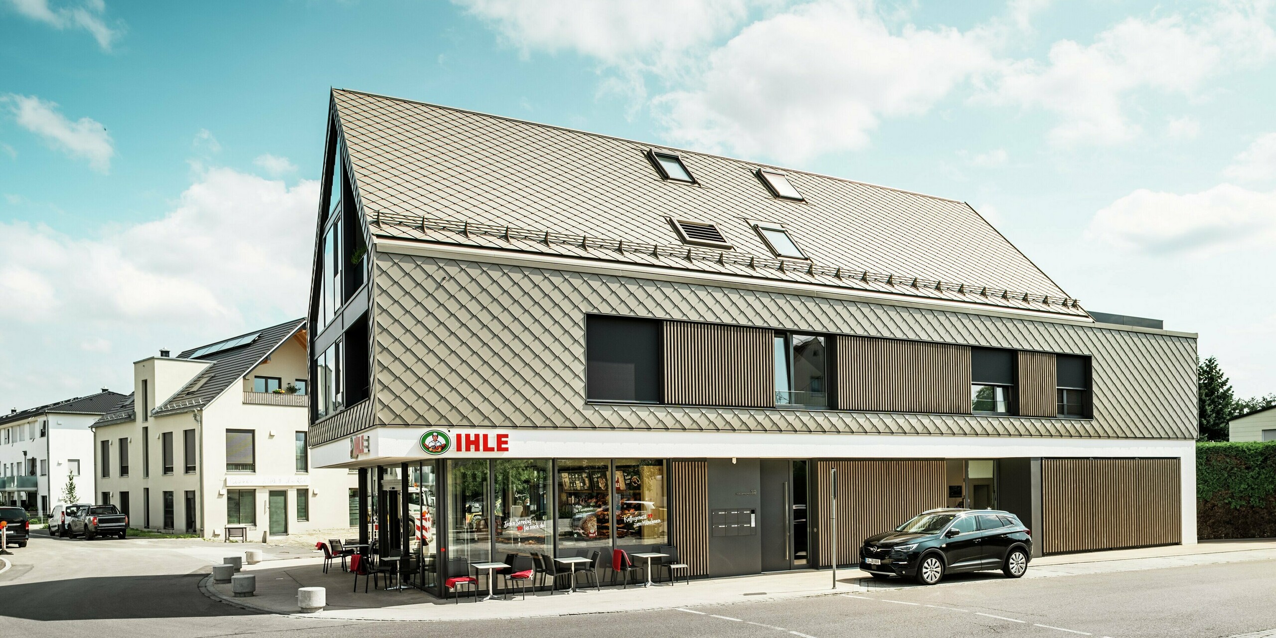 Immeuble d'appartements à Leitershofen, en Allemagne, avec espaces de restauration intégrés au rez-de-chaussée. Le bâtiment à l'architecture sophistiquée a été recouvert de losanges de toiture et de façade 29x29 PREFA élégants en bronze. Les losanges en aluminium confèrent au bâtiment une coque élégante et résistante aux intempéries. De grandes surfaces vitrées et des éléments en bois soulignent le design sophistiqué, tandis que la façade en plâtre blanc du rez-de-chaussée crée un contraste clair.