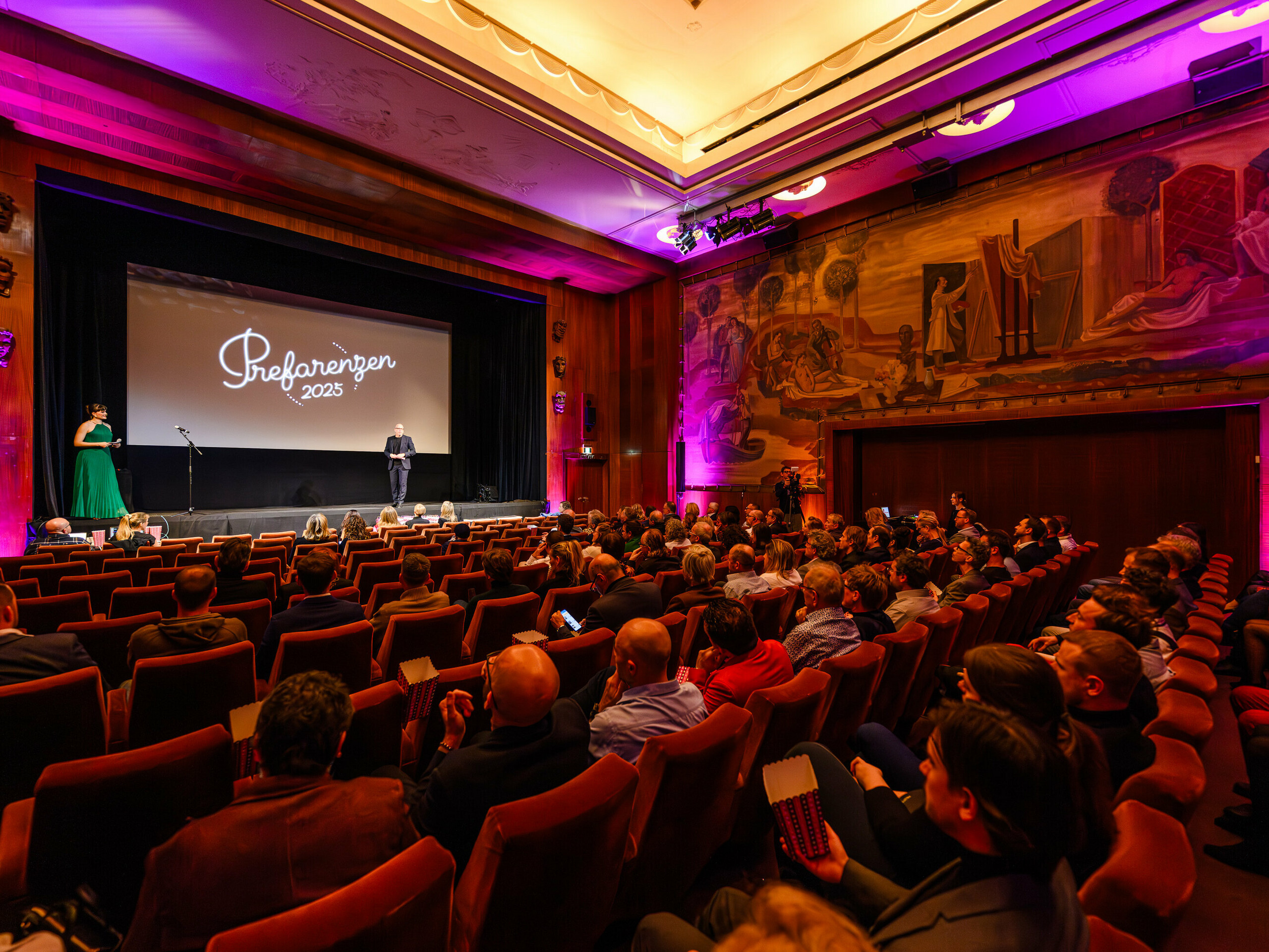 Le cinéma municipal historique de Vienne lors de la première édition des PREFARENZEN 2025 : Jürgen Jungmair, directeur marketing de PREFA, s’adresse à un public international d’experts en architecture, urbanisme et médias. À l’arrière-plan, le logo « PREFARENZEN 2025 » s’affiche sur l’écran, encadré par un éclairage élégant et une œuvre murale impressionnante.