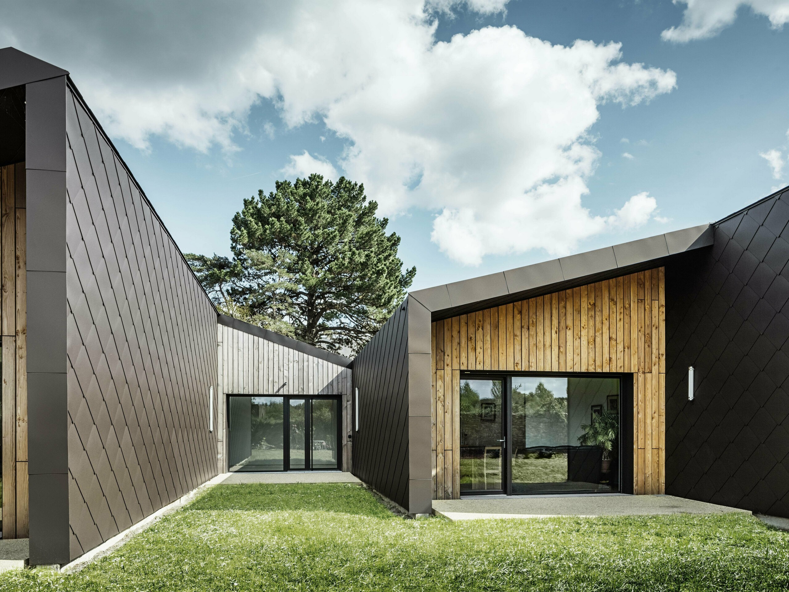 La photo montre une architecture moderne avec une façade en losanges de toiture et de façade PREFA 44 × 44 de couleur brun noisette. La structure en losange du revêtement en aluminium confère au bâtiment un aspect élégant et géométrique. La façade combine harmonieusement des éléments en bois naturel et de grandes surfaces vitrées, créant une atmosphère chaleureuse et accueillante. La pelouse verte et le ciel dégagé renforcent l'impression à la fois moderne et proche de la nature de l'architecture.