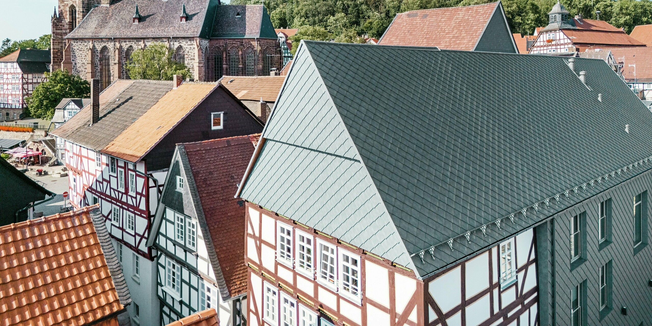 Le toit de la Volkshaus rénovée à Homberg, DE, avec des losanges 29x29 en aluminium PREFAen P.10 gris pierre, intégré dans la vieille ville historique avec des maisons à colombages, une église et un château. Le revêtement en aluminium durable et robuste allie un design moderne au charme de l'environnement historique. Au centre de l'image, on voit le pignon du bâtiment, à l'arrière-plan l'église et la colline du château.