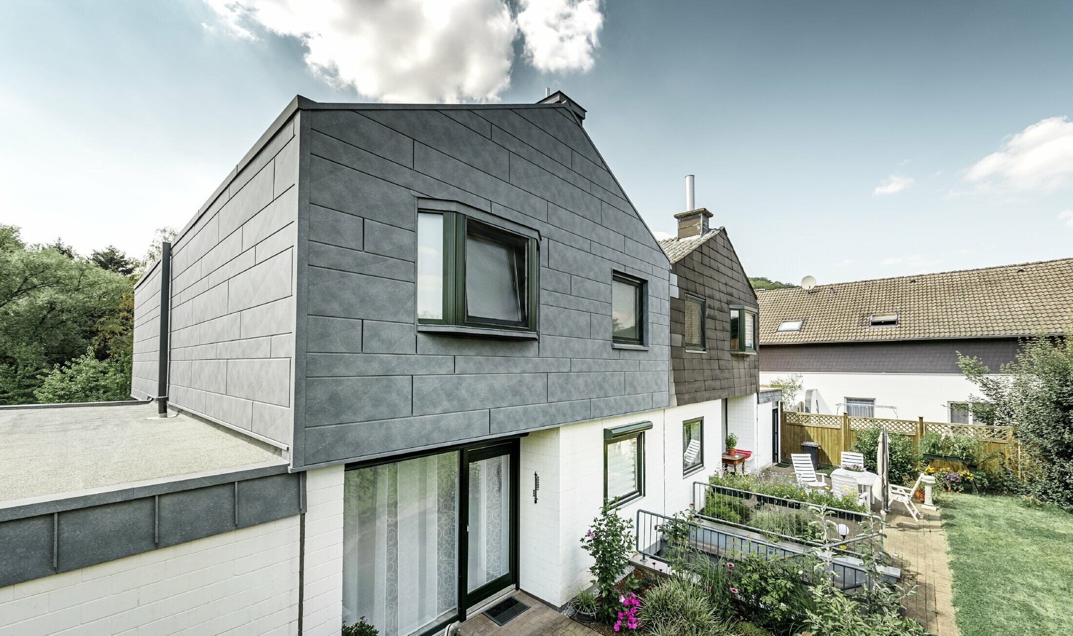 La partie haute du bâtiment de la moitié de maison jumelée a été rénovée avec l’élément de façade Siding.X PREFA en P.10 gris pierre. Juste à côté pour la comparaison, la moitié de maison avant la rénovation.