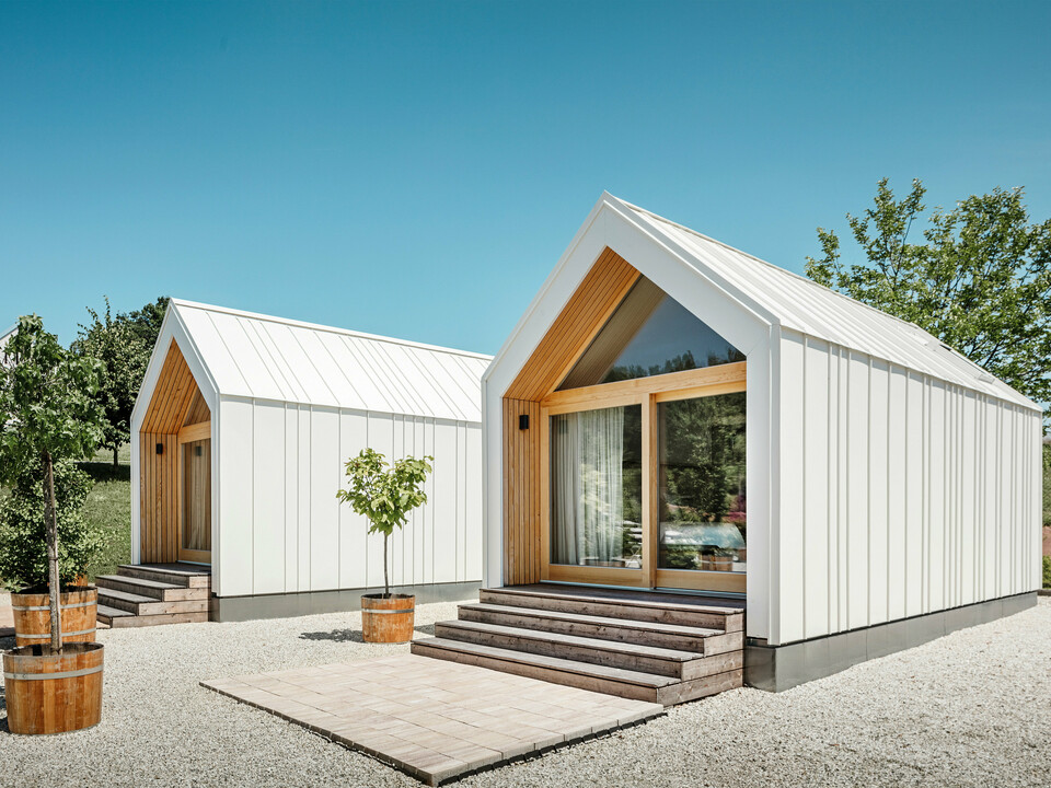 Deux petites maisons blanches dans l'idylle de vacances « Pri Momi » à Kančevci, en Slovénie, conçues avec un revêtement de toit et de façade PREFALZ en blanc pur P.10 et en bois naturel. L'architecture minimaliste des bâtiments est soulignée par l'élégant mélange de matériaux aluminium et bois. Les grandes façades vitrées aux entrées ainsi que les marches en bois et les arbres environnants créent une atmosphère invitante et harmonieuse. L'hébergement exclusif est situé au milieu d'un paysage pittoresque et est utilisé spécifiquement pour les célébrations.