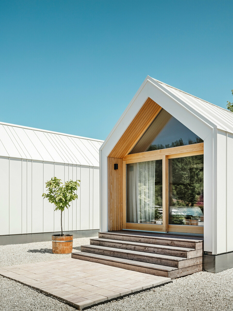 Deux petites maisons blanches dans l'idylle de vacances « Pri Momi » à Kančevci, en Slovénie, conçues avec un revêtement de toit et de façade PREFALZ en blanc pur P.10 et en bois naturel. L'architecture minimaliste des bâtiments est soulignée par l'élégant mélange de matériaux aluminium et bois. Les grandes façades vitrées aux entrées ainsi que les marches en bois et les arbres environnants créent une atmosphère invitante et harmonieuse. L'hébergement exclusif est situé au milieu d'un paysage pittoresque et est utilisé spécifiquement pour les célébrations.